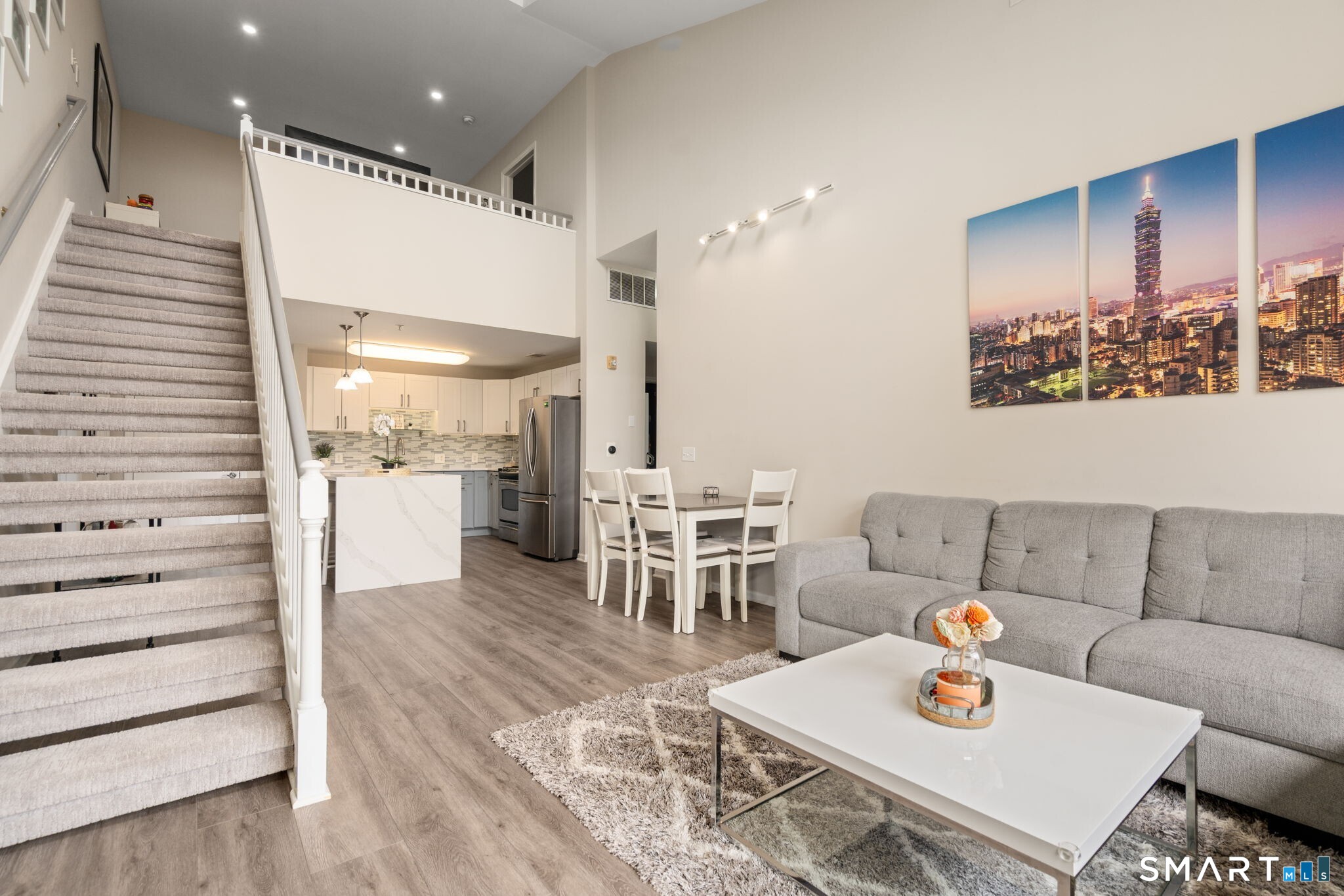 Open-plan living room with gray sofa, white coffee table, kitchen, and carpeted staircase.