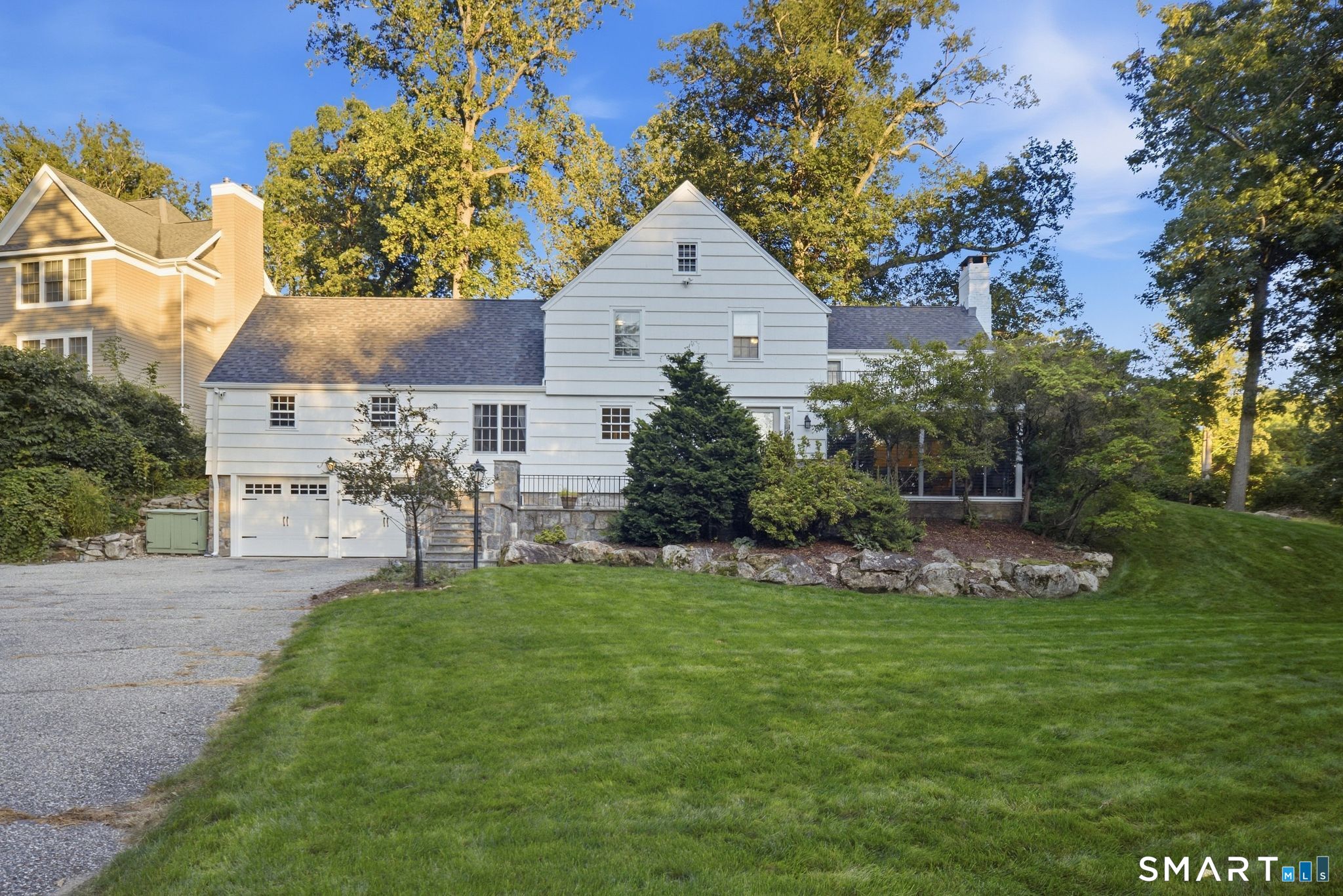 White clapboard house with a central gable, attached garage, mature trees, and a broad green lawn.