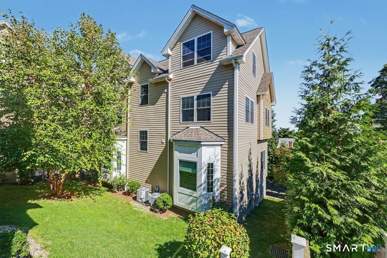 Tan multi-story house with a projecting bay window, surrounded by trees and lawn.