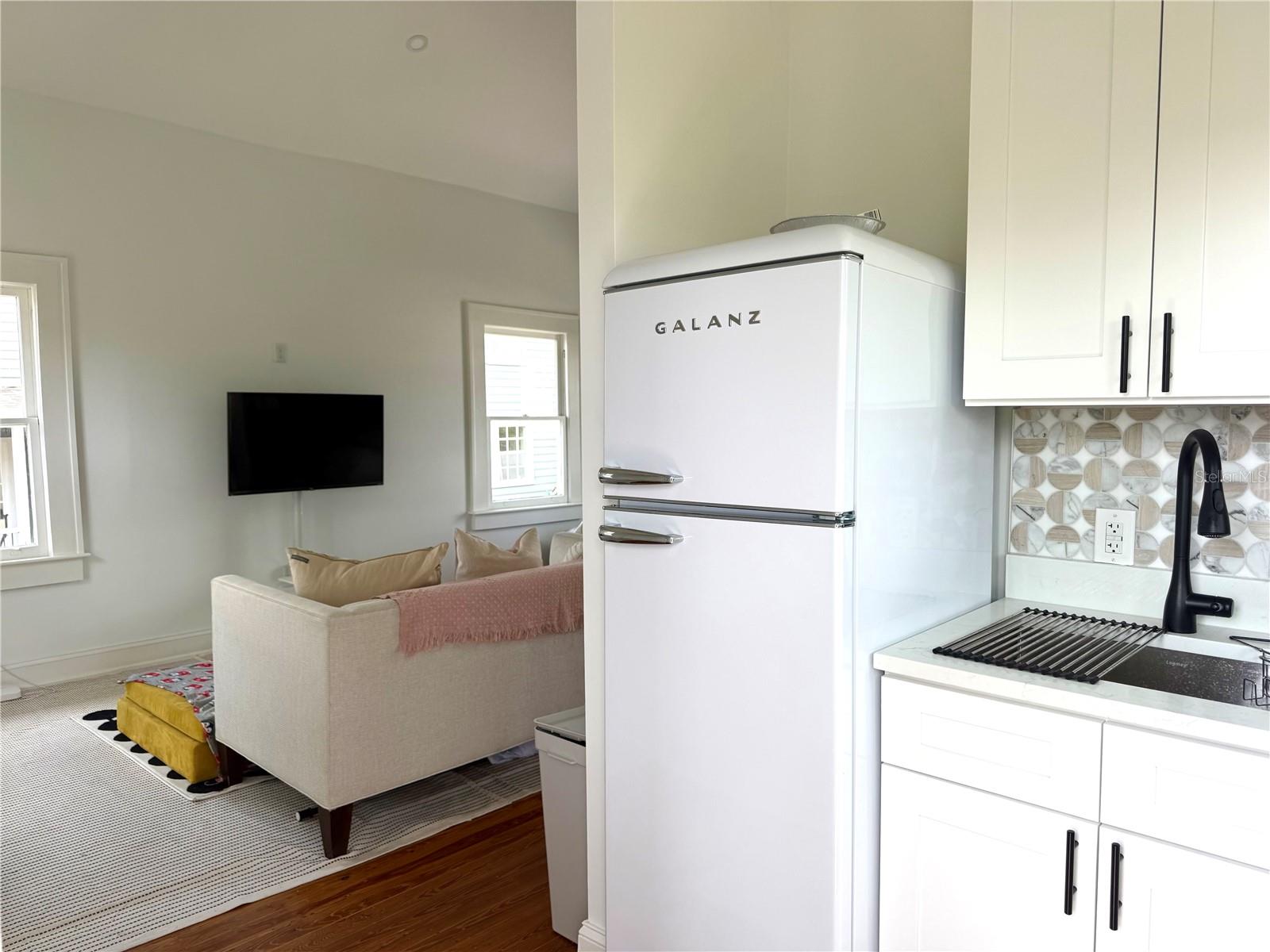 Kitchen with a white GALANZ refrigerator opening onto a living area with a beige sofa and wall-mounted TV.