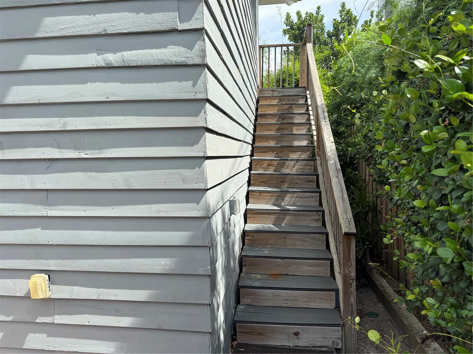 Wooden exterior stairs beside gray house siding, with a handrail and dense greenery.