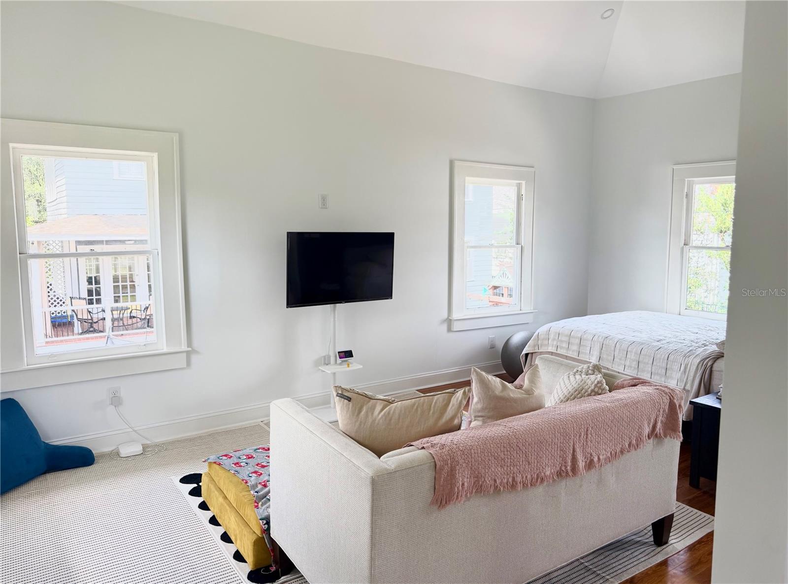 Bright living room with wall-mounted television, large windows, and a white sofa beside a bed.