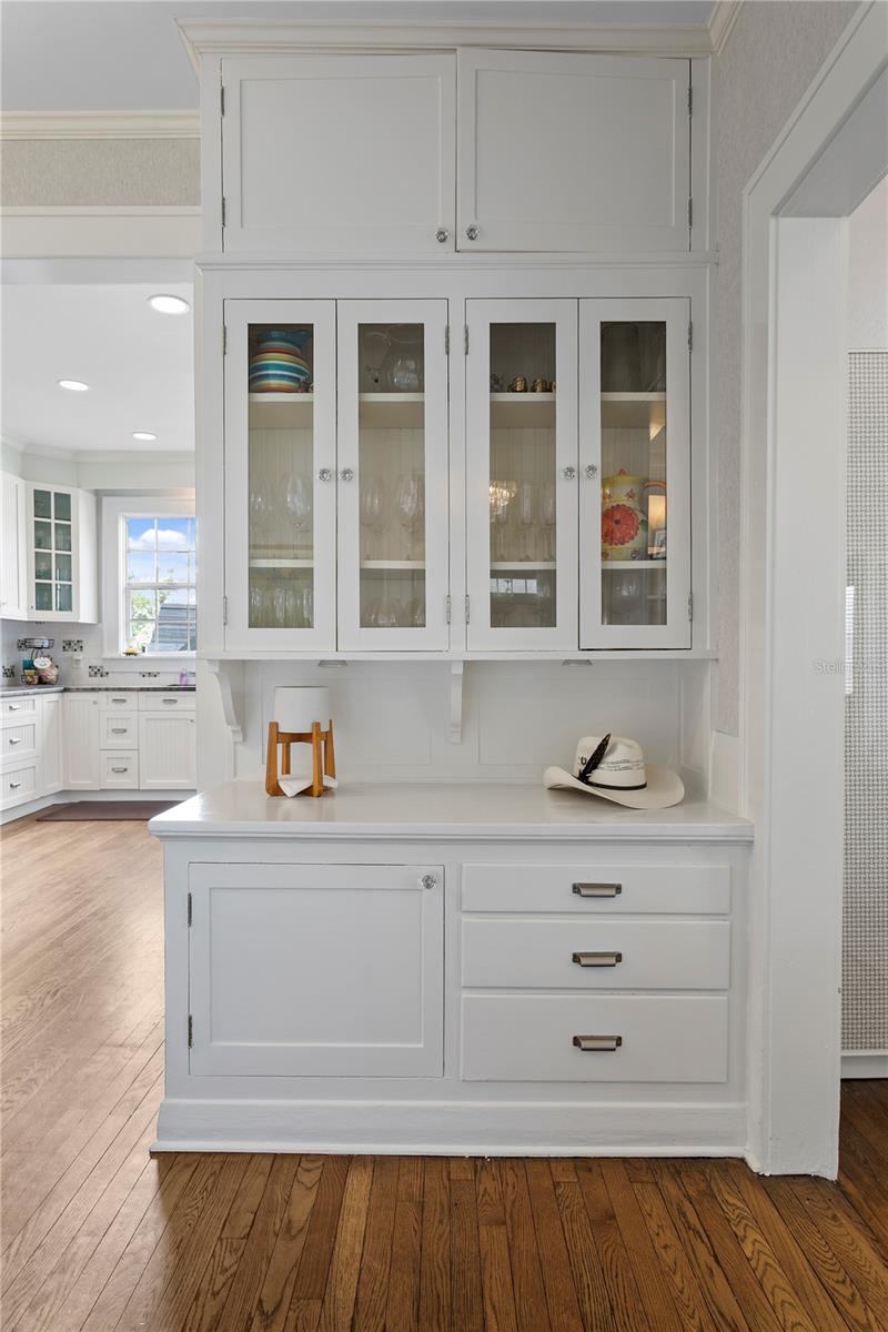 White kitchen built-in with glass-front cabinets, displayed dishes, a counter, and hardwood floors.