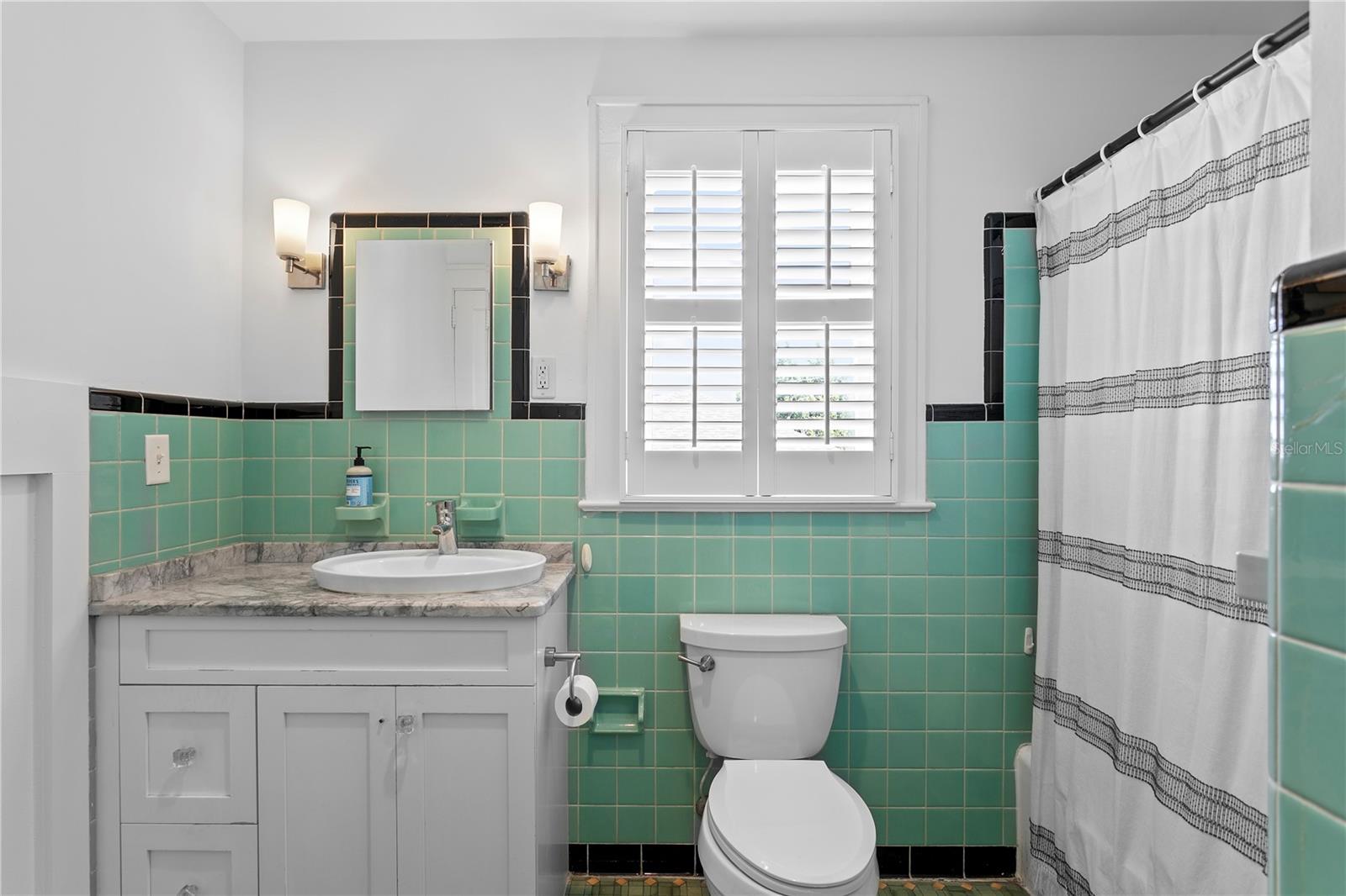 Bathroom with mint-green tile, marble vanity countertop, shuttered window, and white shower curtain.
