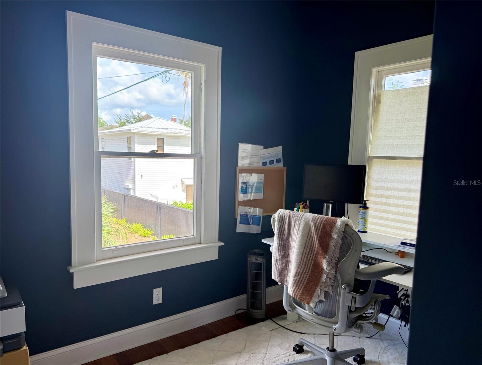 Home office with deep blue walls, two bright windows, and a desk beside a chair draped with a striped blanket.