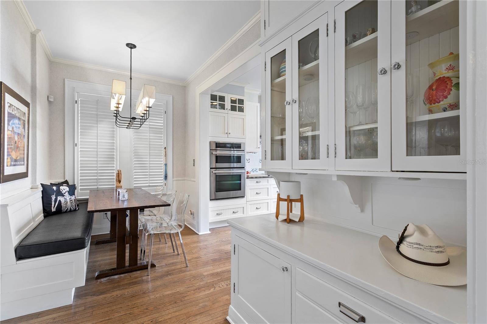 White kitchen with glass-front cabinets and double oven beside a dining nook with a wood table and bench.