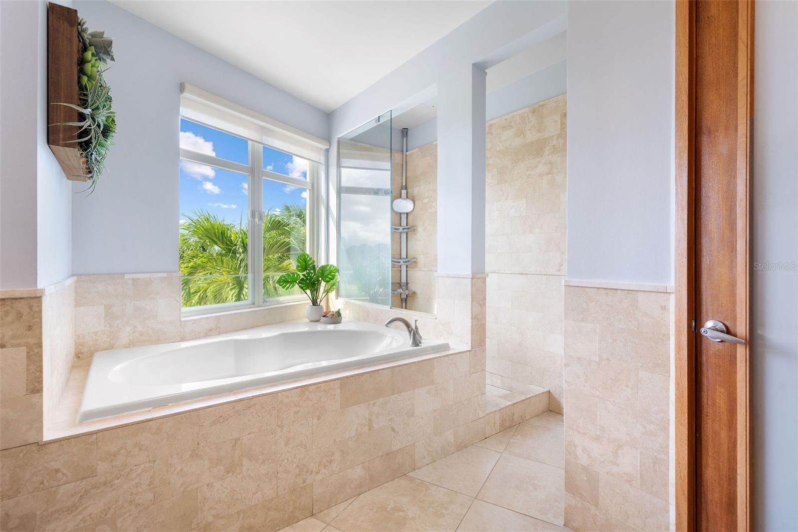 Bathroom with a built-in soaking tub, glass shower, and window overlooking palm foliage.