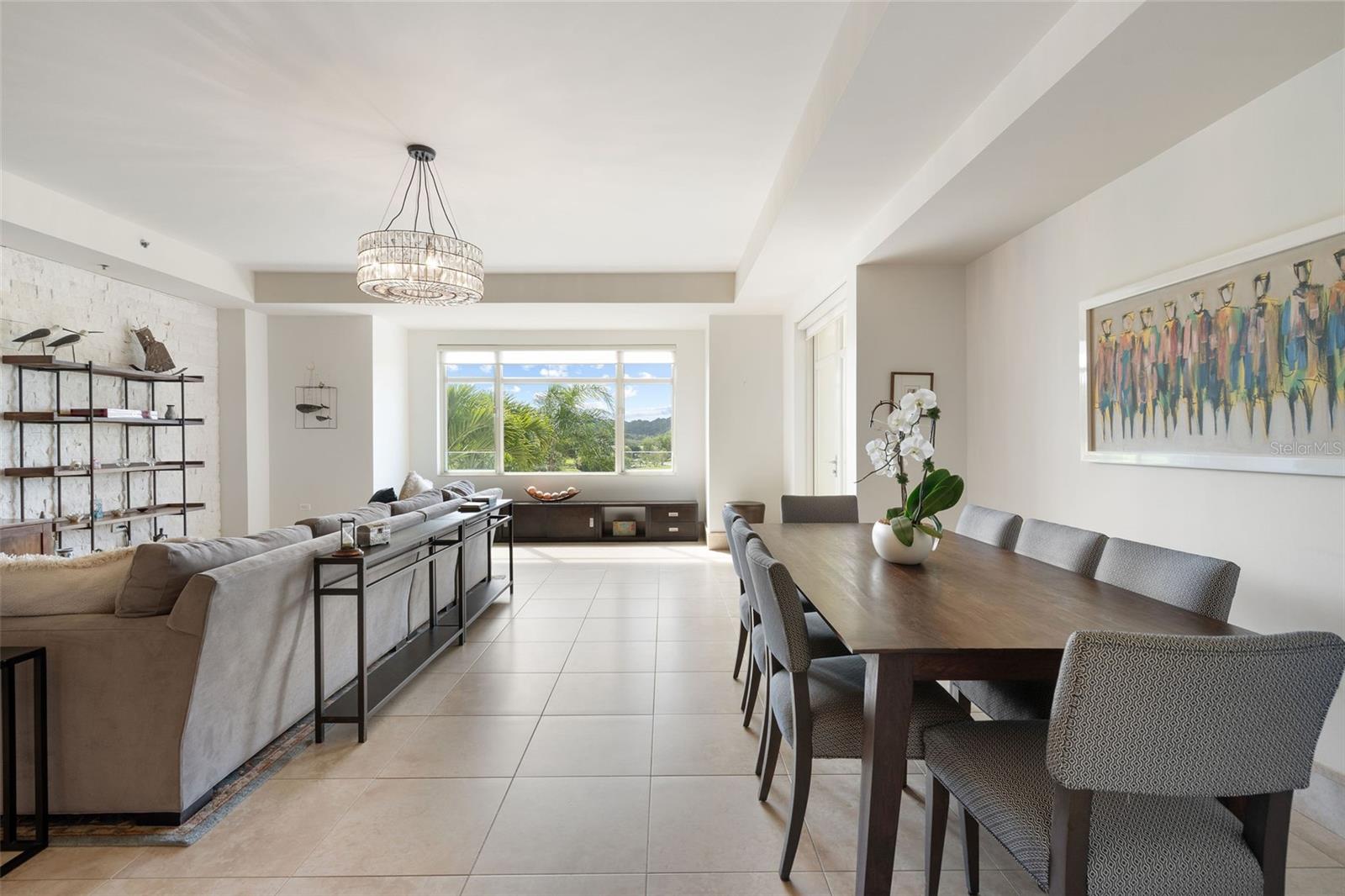 Open-plan living and dining room with a stone accent wall, long wood table, and windows overlooking palm trees.
