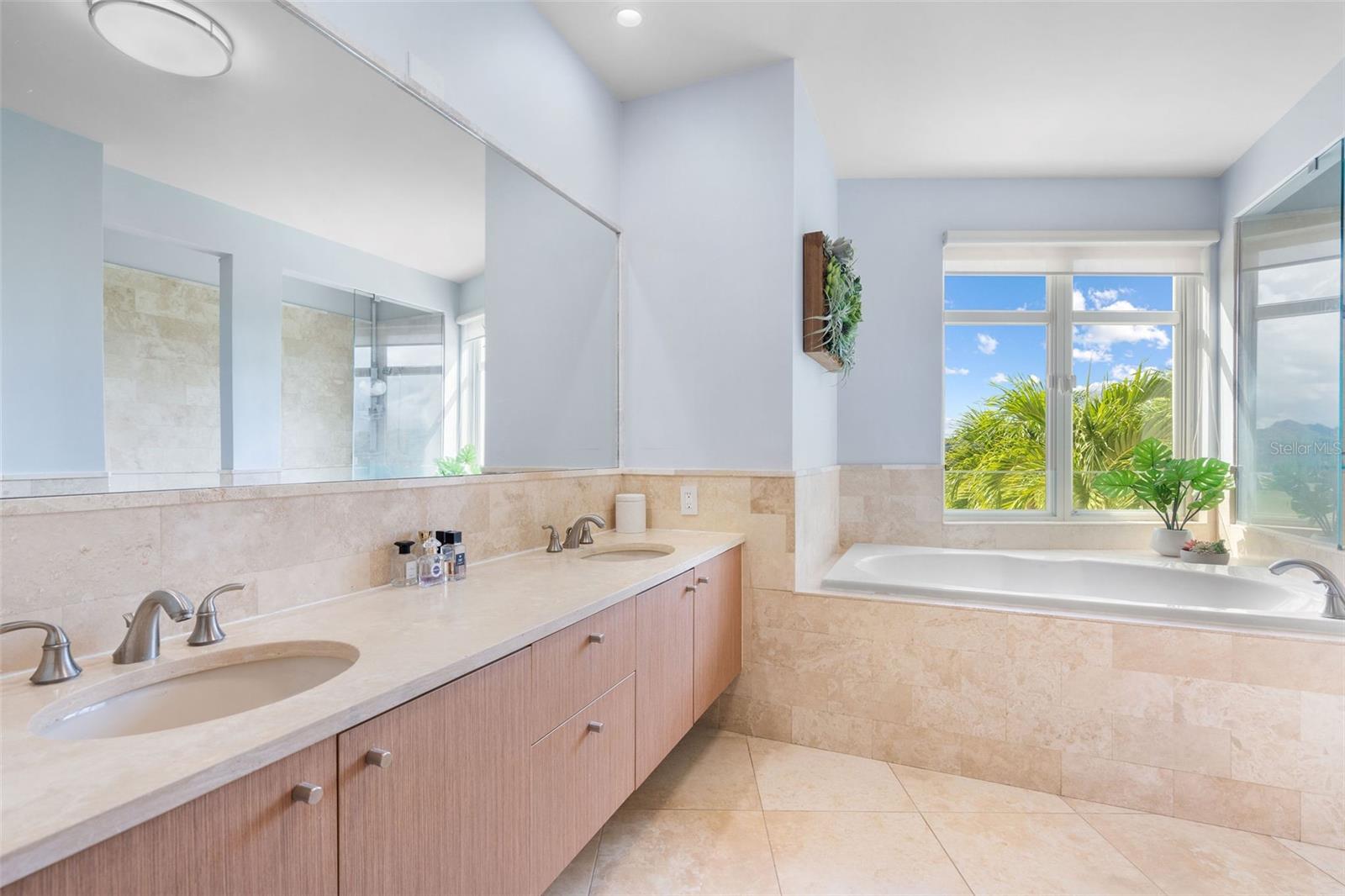 Bathroom with a double-sink vanity and a built-in tub beside windows overlooking tropical greenery.