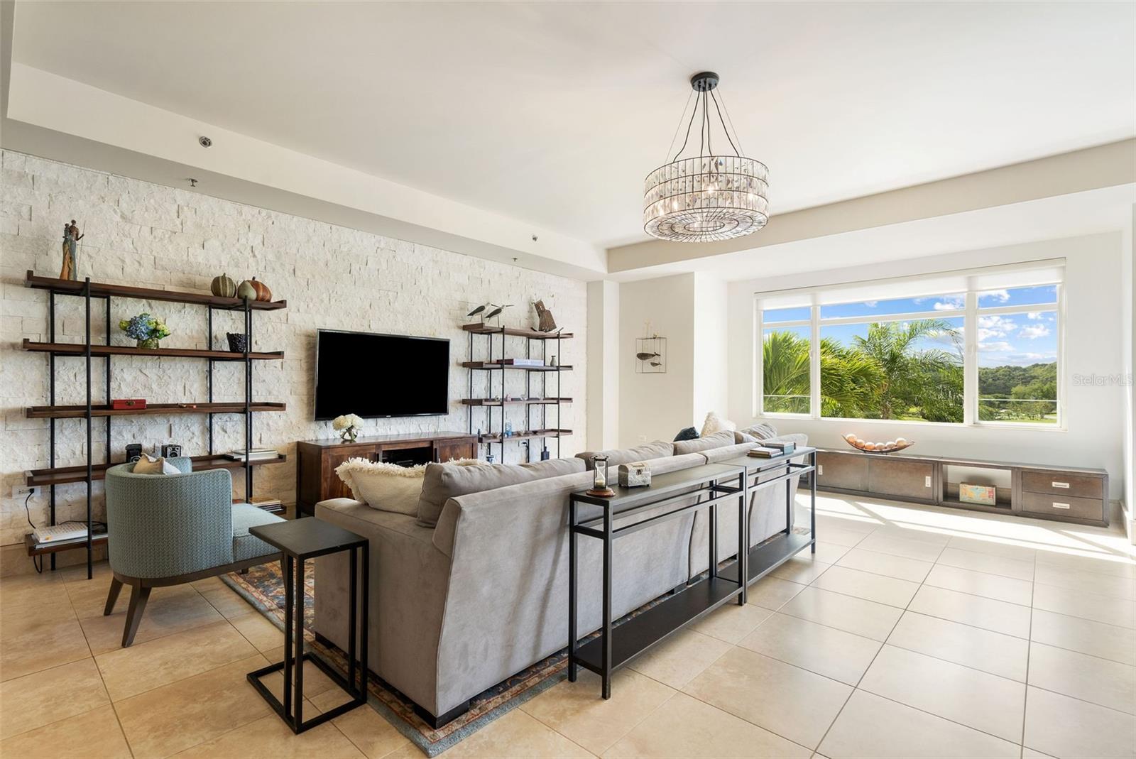 Furnished living room with a stone accent wall, chandelier, and large windows overlooking palm trees.