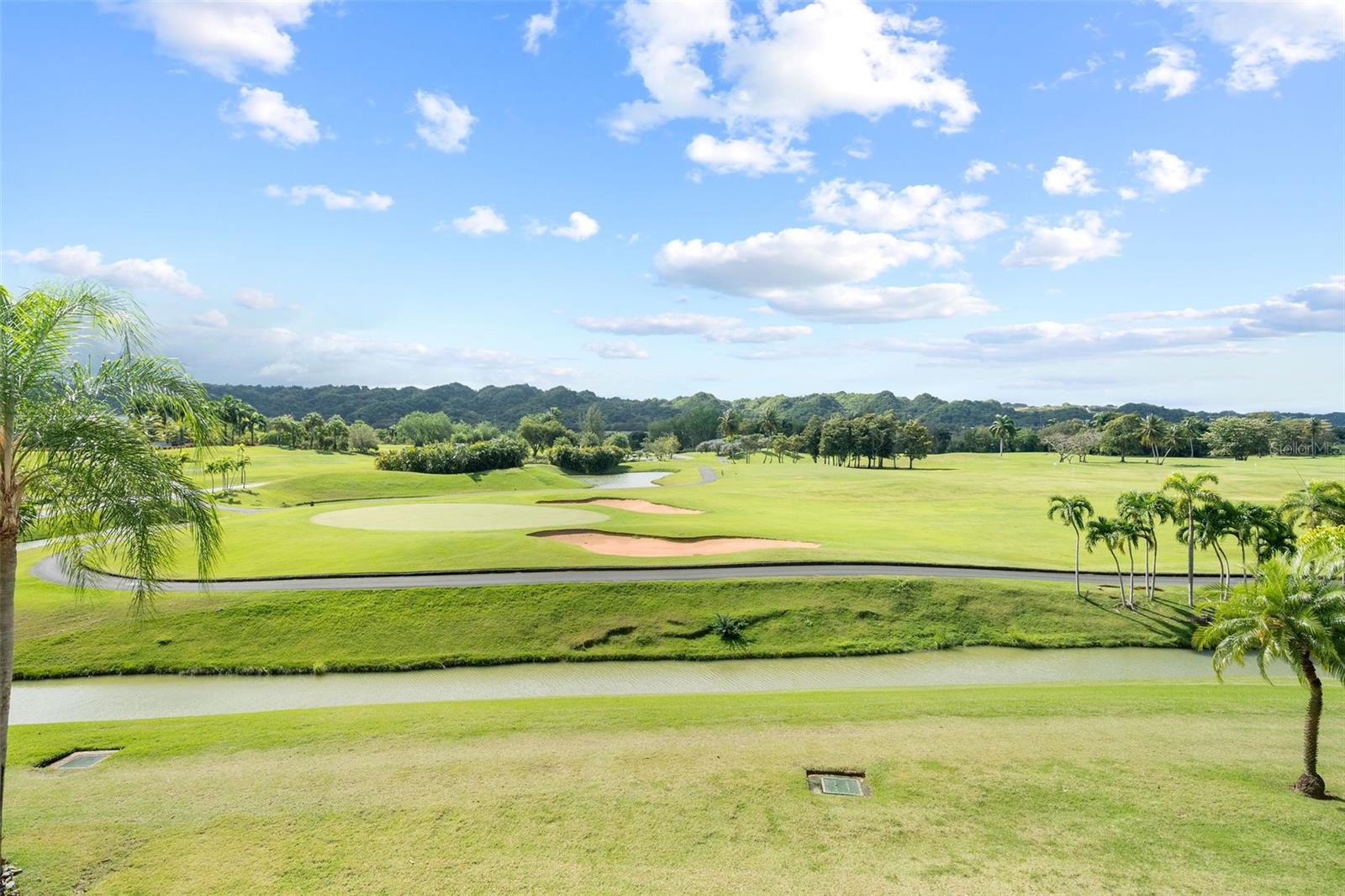 Golf course view with sand bunkers, palm trees, a foreground water channel, and wooded hills.