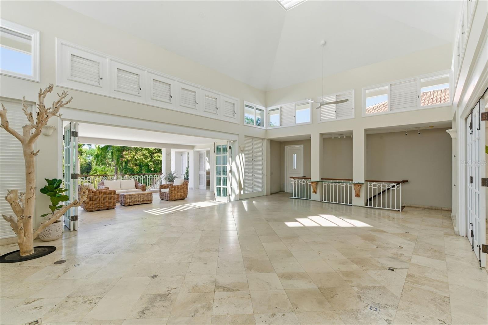 Double-height living area with pale stone floors, louvered windows, and open doors to a covered terrace.