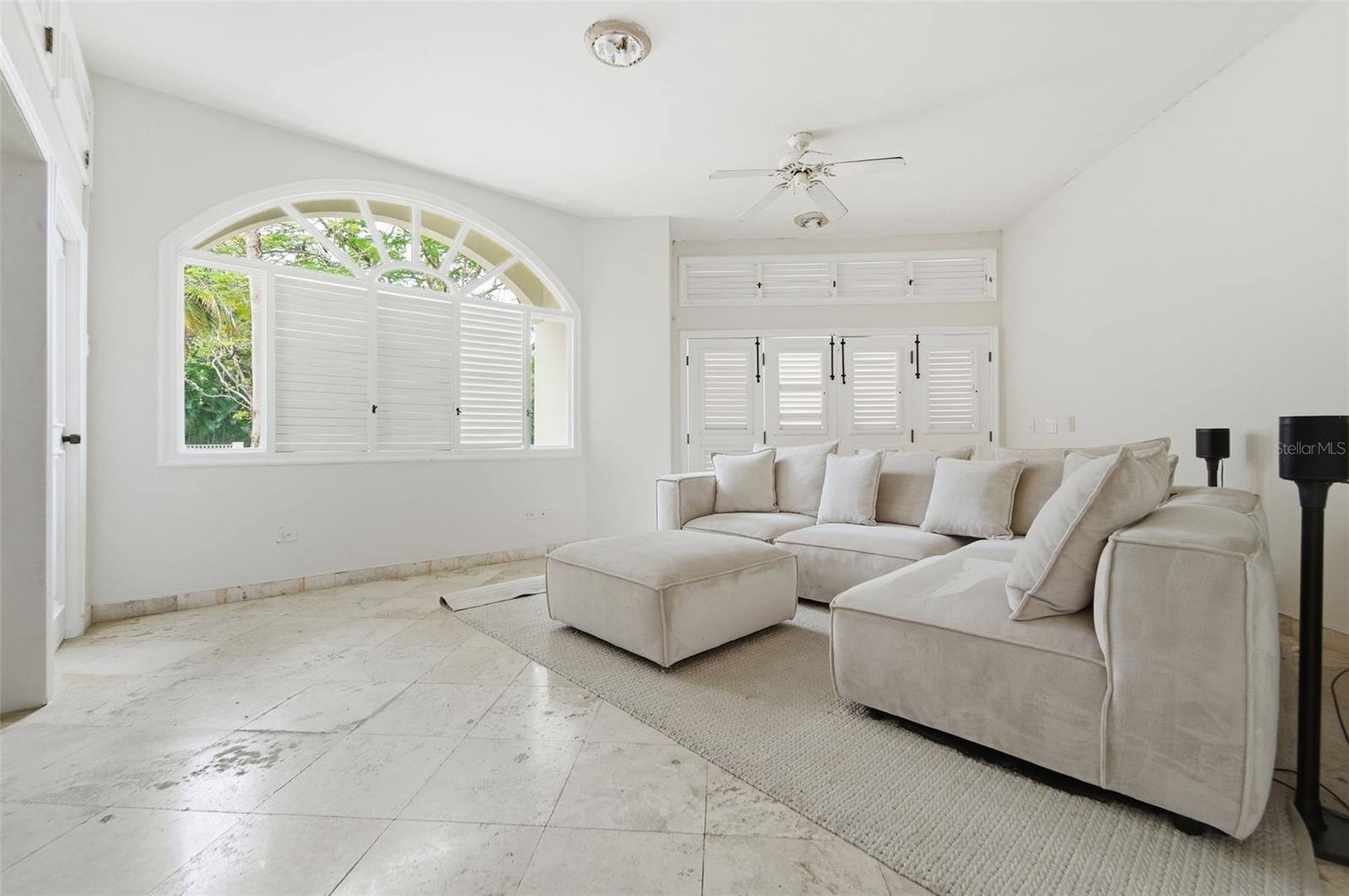 Living room with a cream sectional, stone tile flooring, and a large arched shuttered window.