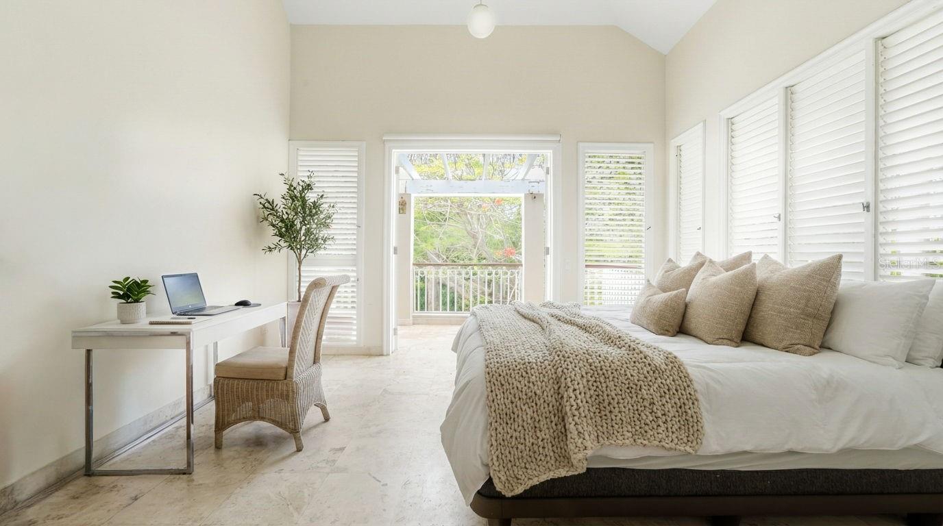 Furnished bedroom with a white bed, knitted throw, desk, and balcony overlooking greenery.