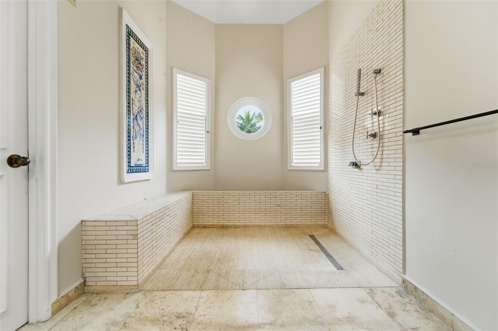 Bathroom with a tiled walk-in shower, built-in bench, and round window overlooking palm trees.