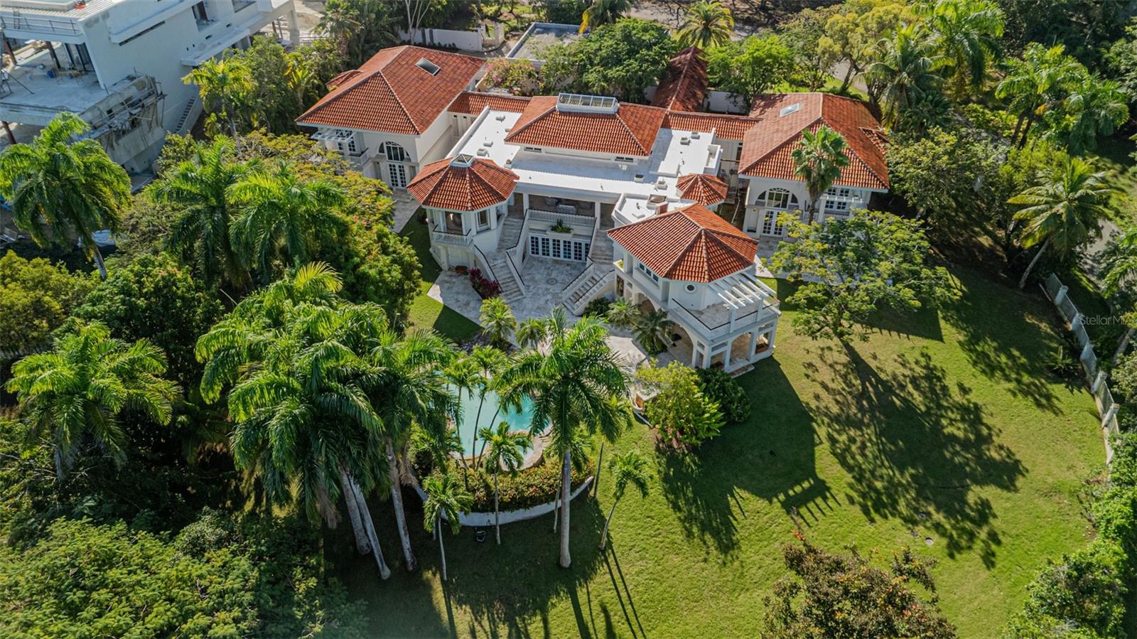 Aerial view of a white estate with terracotta roofs, tropical landscaping, and a pool.