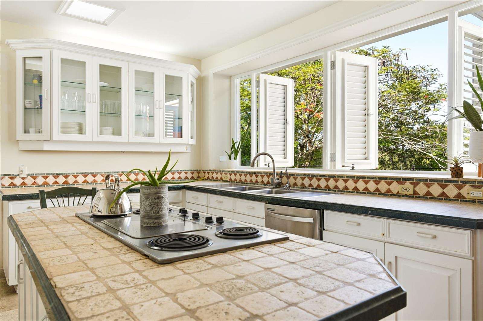 Bright kitchen with a tiled cooktop island and open shuttered windows overlooking trees.