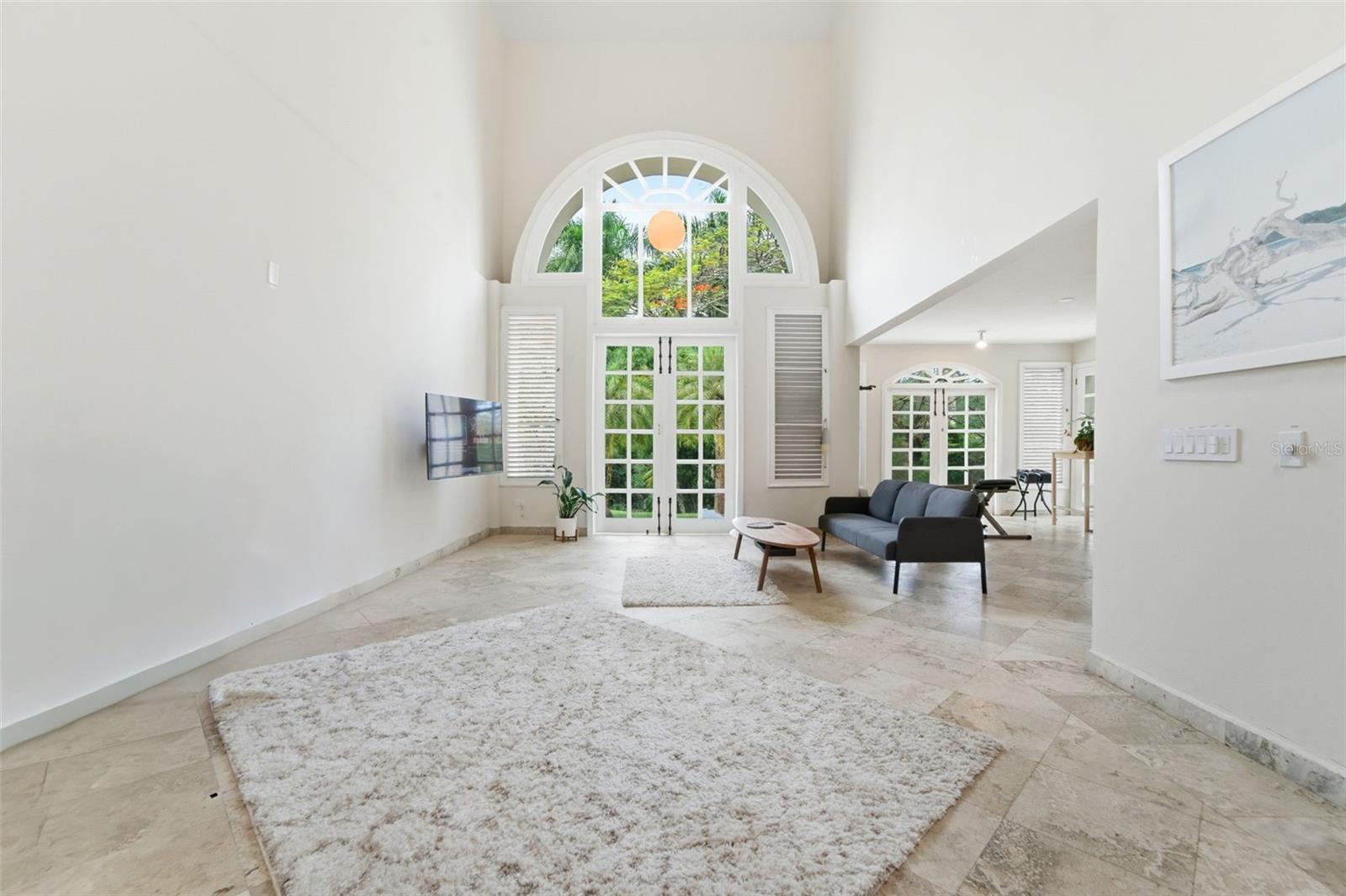 Living room with a soaring arched window, French doors, pale stone floors, and a dark sofa.
