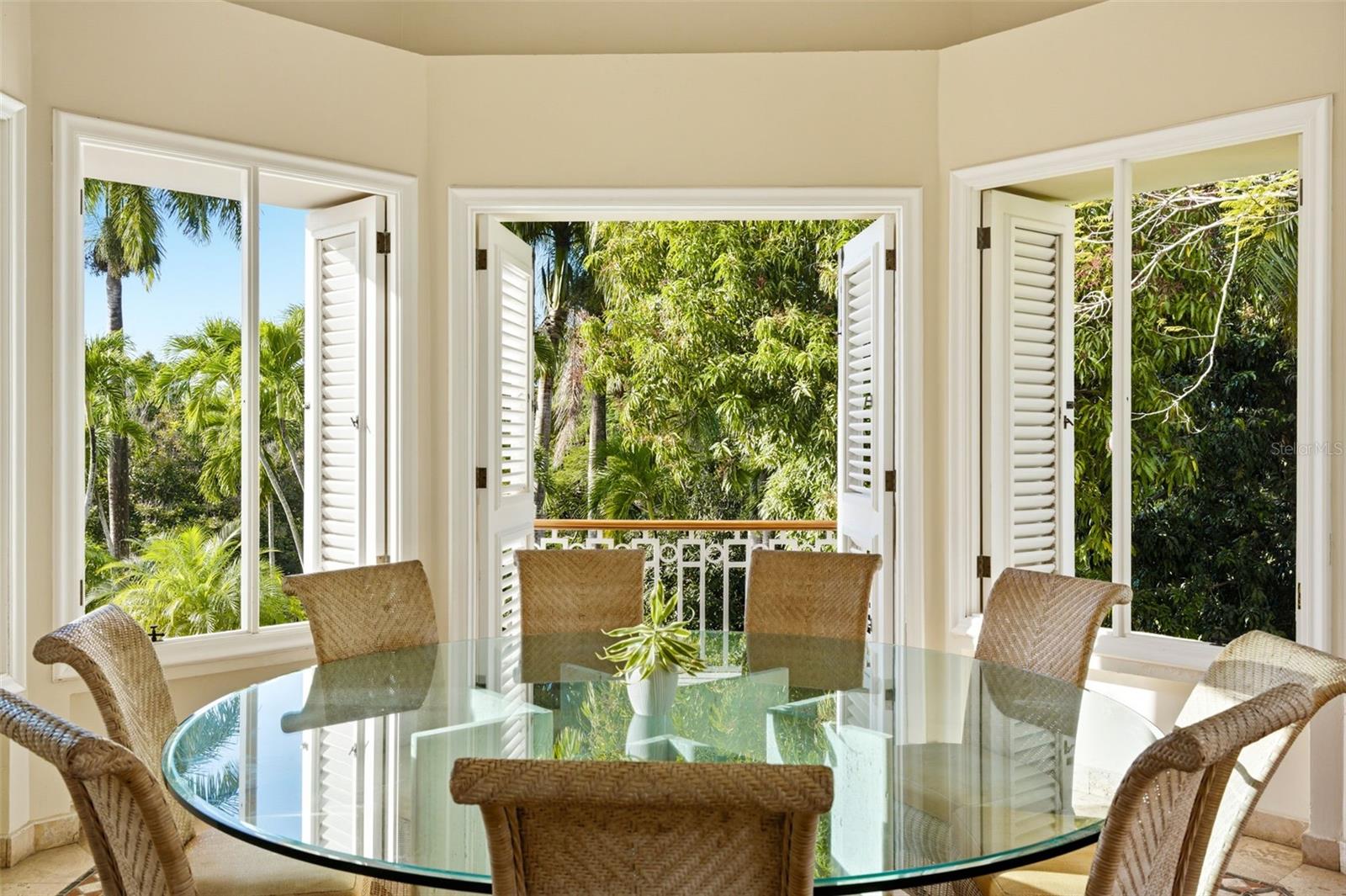 Dining room with a glass table and woven chairs facing open doors and tropical greenery.