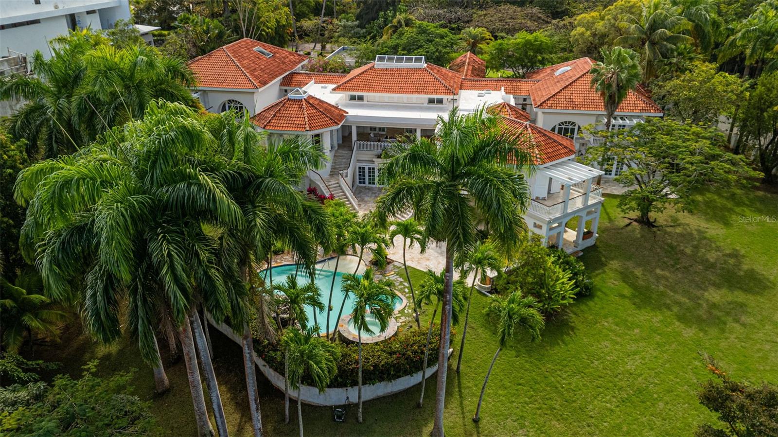 Aerial view of a white estate with red tile roofs, a curved pool, and tropical palms.
