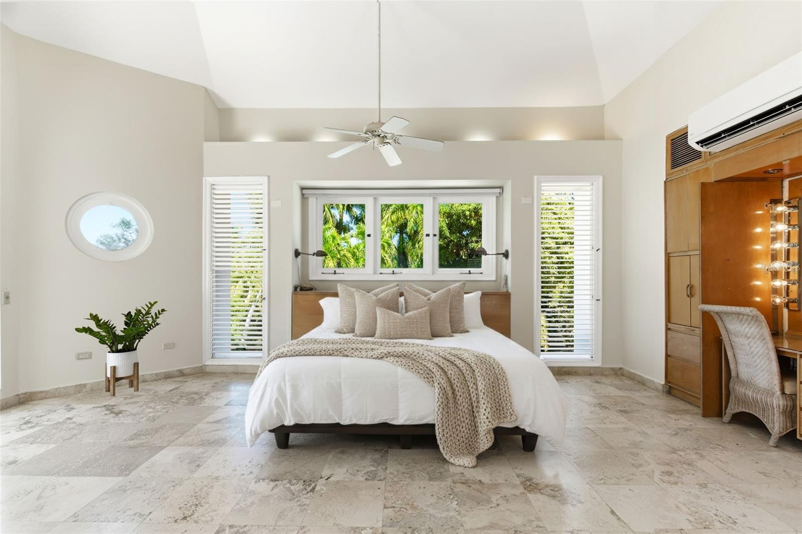 Bedroom with a vaulted ceiling, white bed, tropical greenery outside the windows, and a built-in wood vanity.