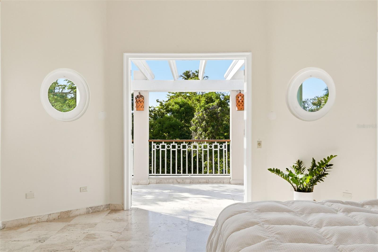 Bright bedroom with marble flooring, round windows, and a balcony opening onto leafy trees.