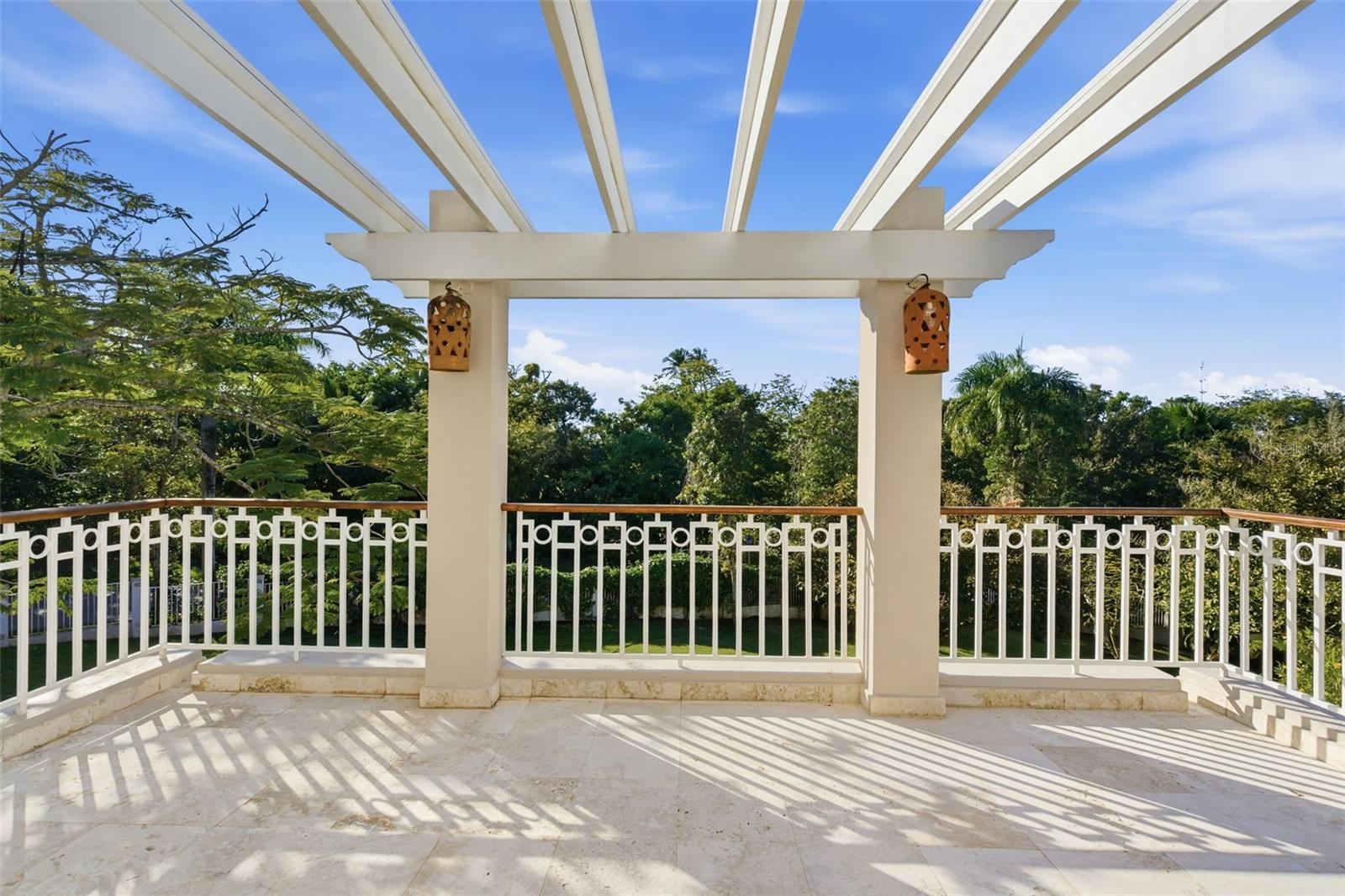 Tiled terrace beneath a white pergola, with geometric railings and tropical trees beyond.