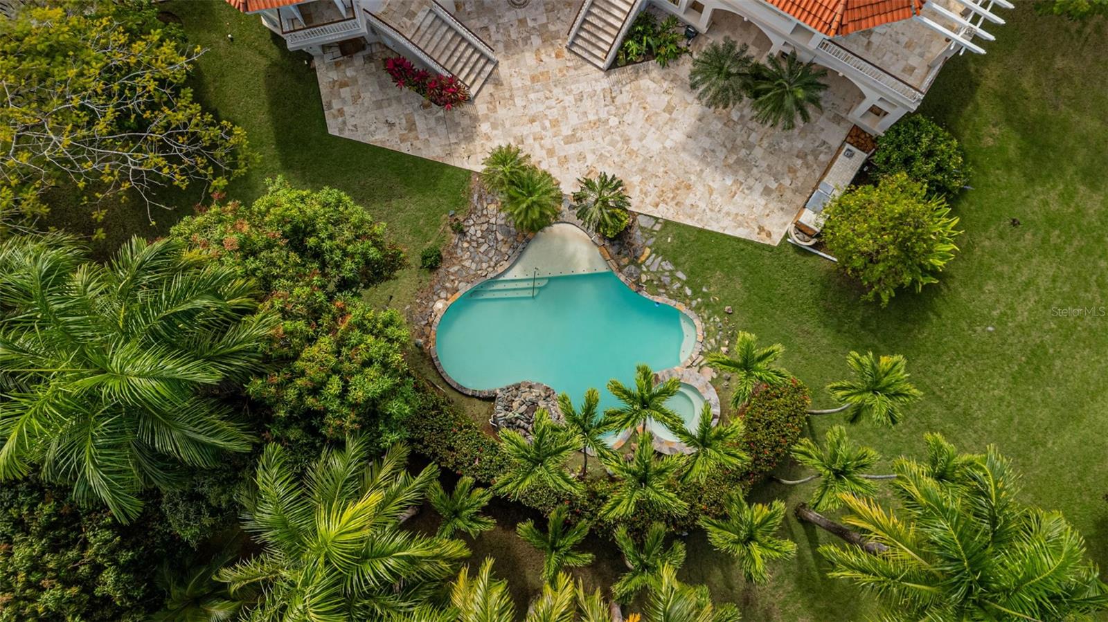 Aerial view of a freeform pool beside a stone patio, terracotta-roofed house, and tropical palms.