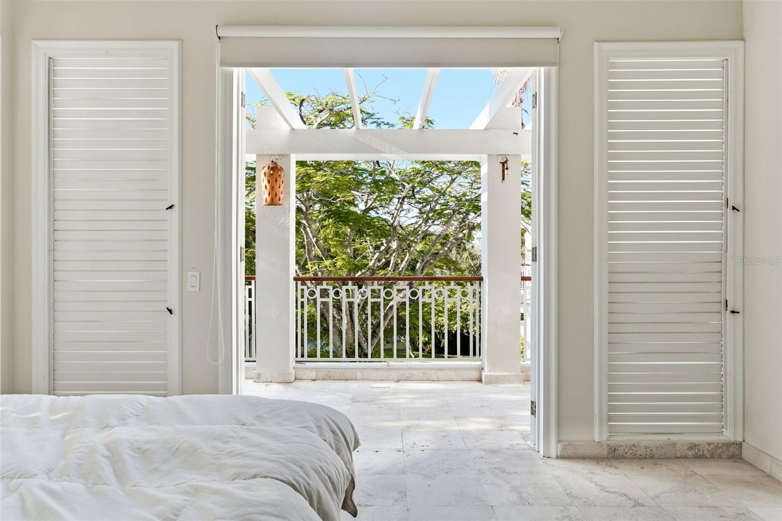 Bedroom opening onto a covered balcony framed by white beams and leafy trees.