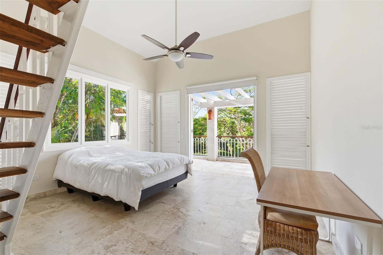 Bedroom with a white bed and staircase, opening onto a greenery-surrounded balcony.