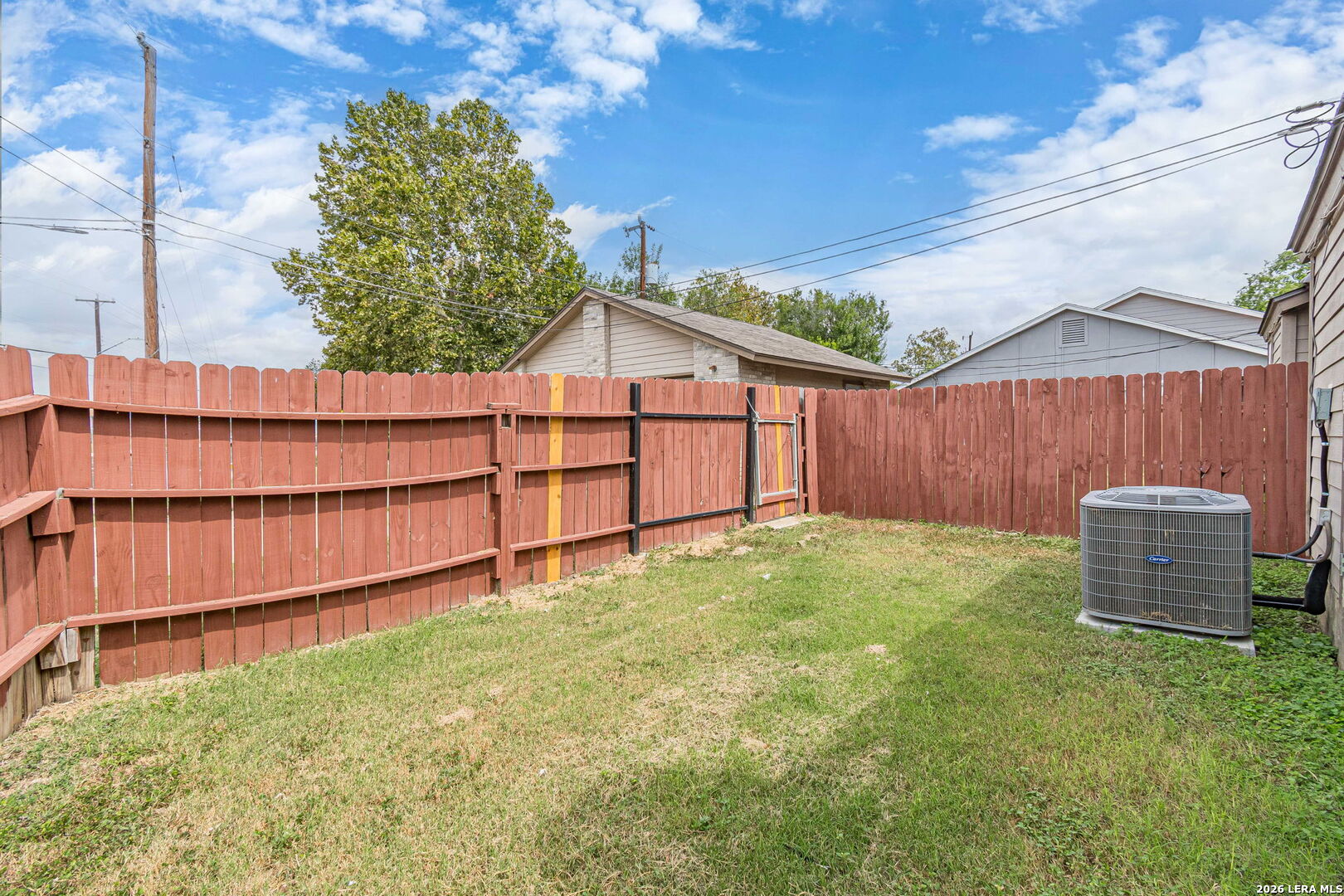 Fenced backyard with a red wooden privacy fence, patchy lawn, and outdoor air-conditioning unit.