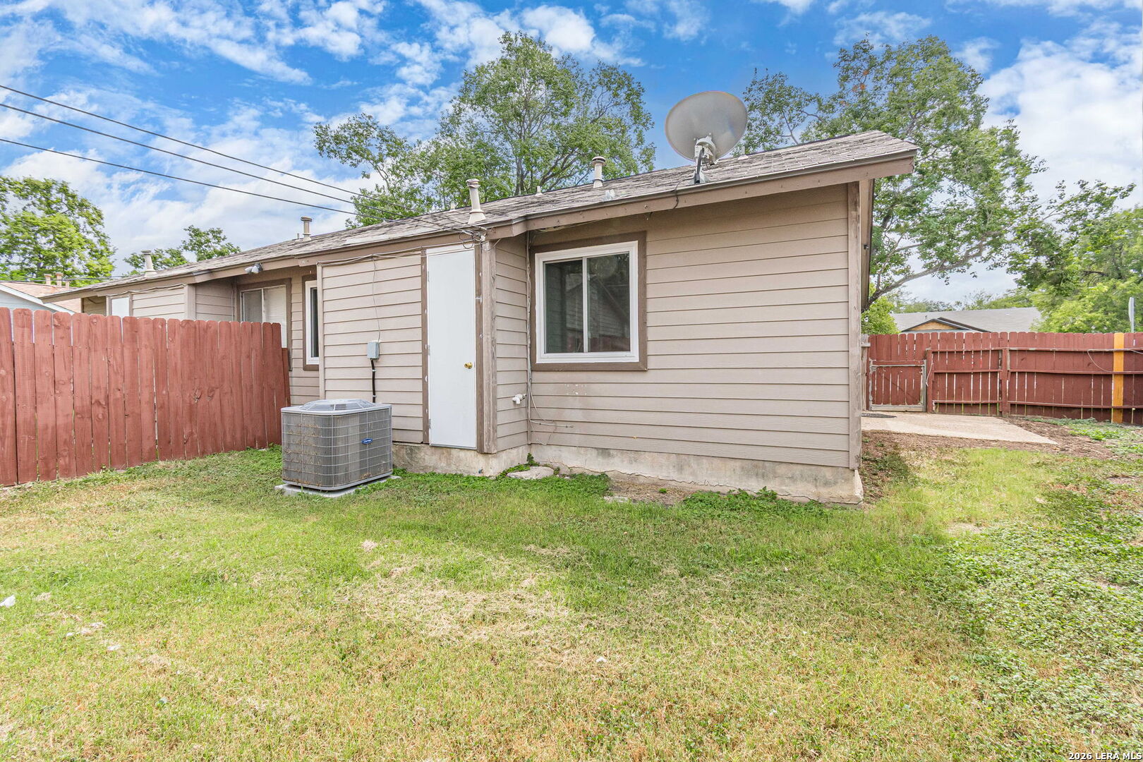 Rear of a tan-sided home with a fenced yard, satellite dish, and outdoor air-conditioning unit.