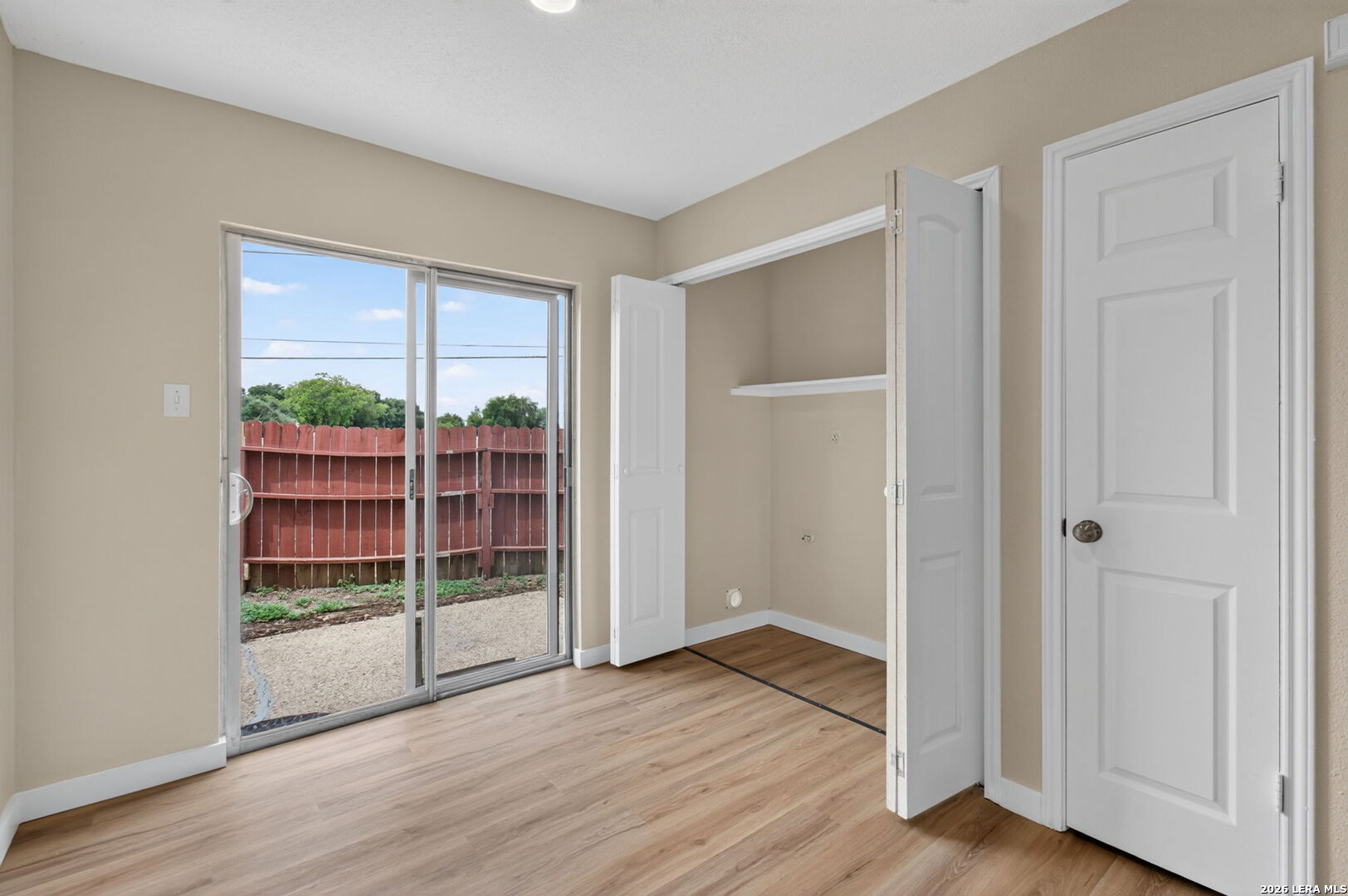 Empty bedroom with light wood flooring, an open closet, and a sliding glass door to a fenced patio.