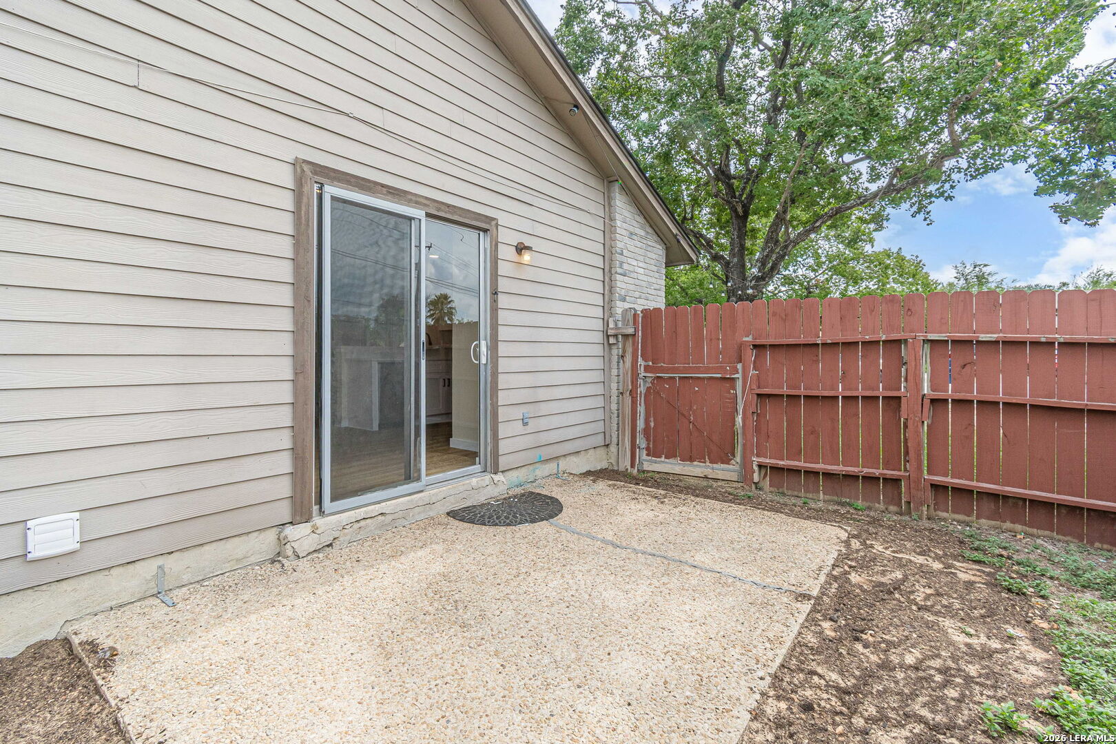 Concrete patio beside a beige-sided house, with a sliding glass door and red wooden privacy fence.