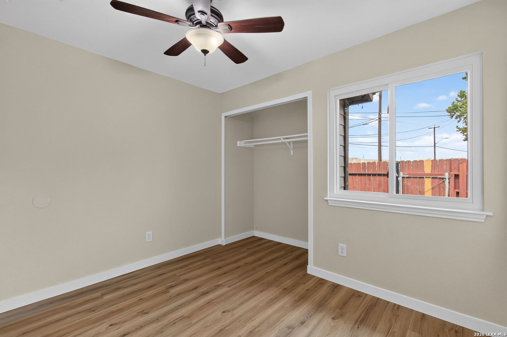 Vacant bedroom with a ceiling fan, open closet, and window overlooking a fenced yard.