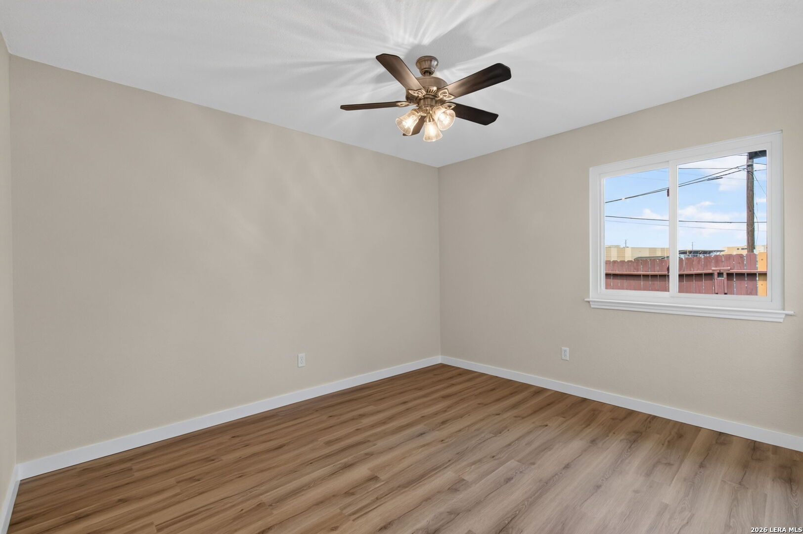 Empty room with wood-look flooring, a lit ceiling fan, and a window overlooking a wooden fence.