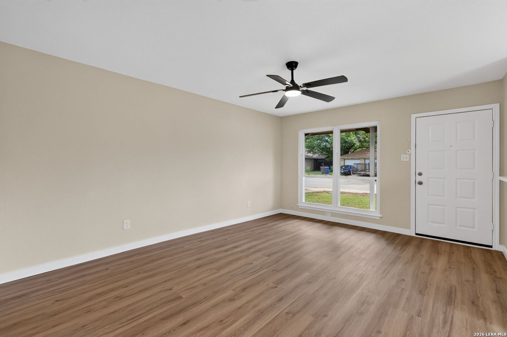 Empty room with wood-look flooring, a ceiling fan, front windows, and a white entry door.