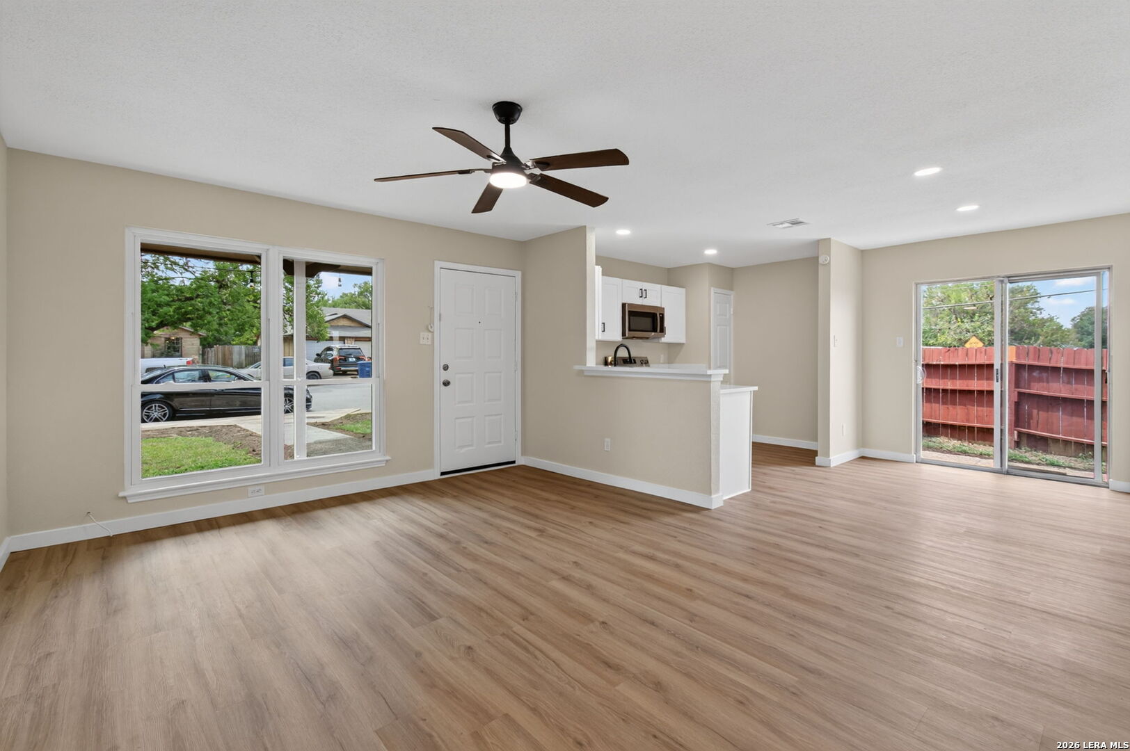 Vacant living room with wood-look floors, ceiling fan, kitchen breakfast bar, and sliding glass doors.