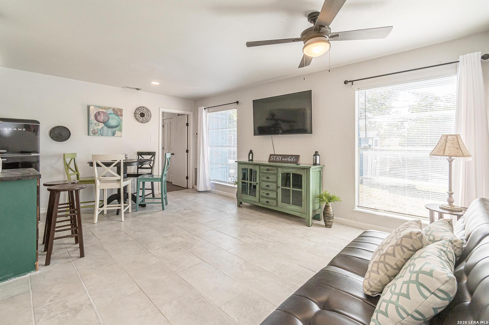 Furnished living and dining area with tiled floors, a green sideboard, wall-mounted TV, and large windows.