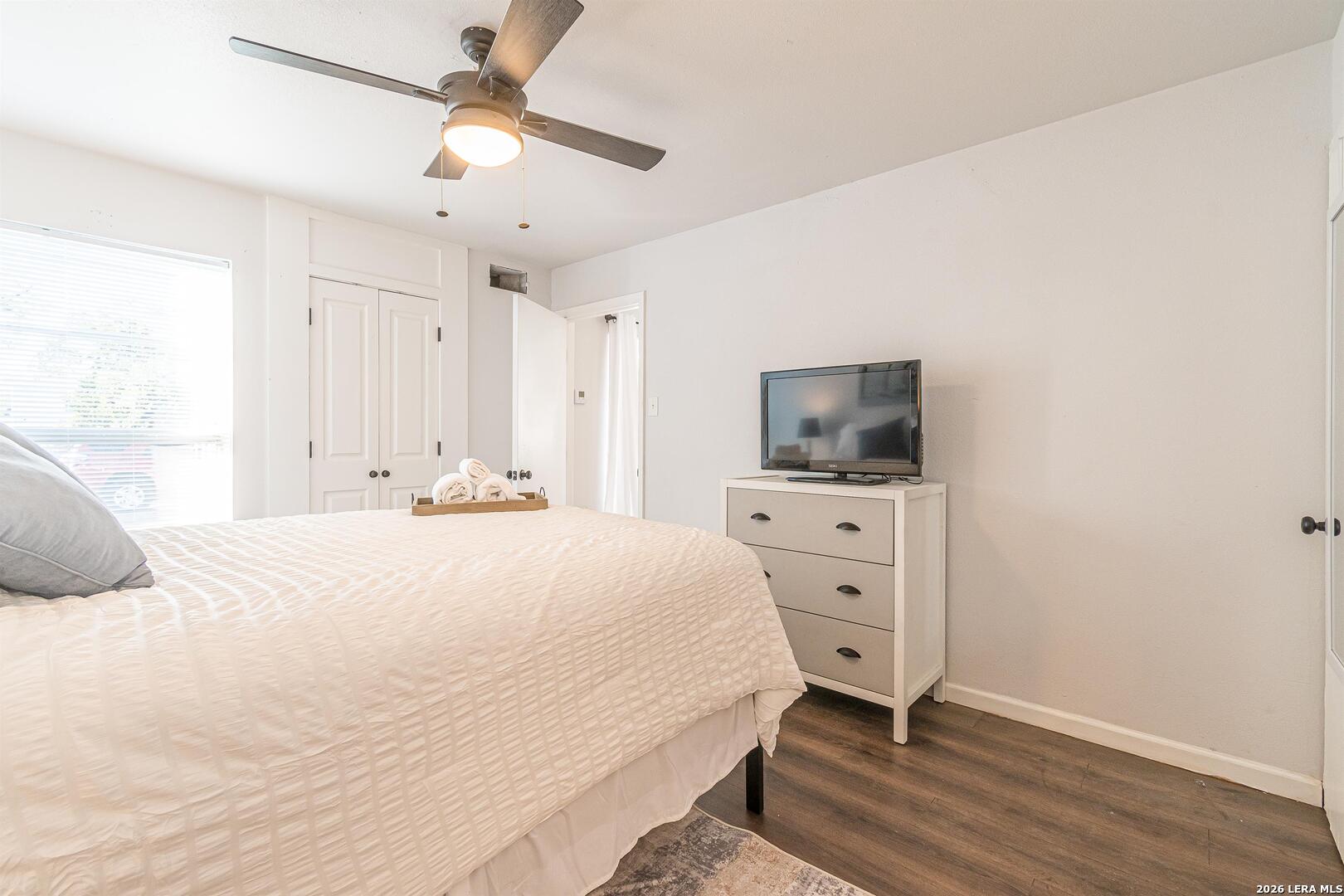 Bedroom with a white quilted bed, window blinds, and a dresser topped with a television.
