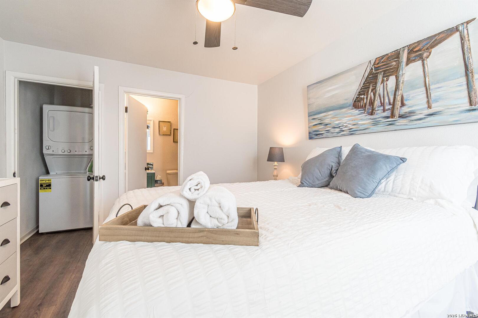 Staged bedroom with a white bed, blue pillows, rolled towels, and an open laundry closet.