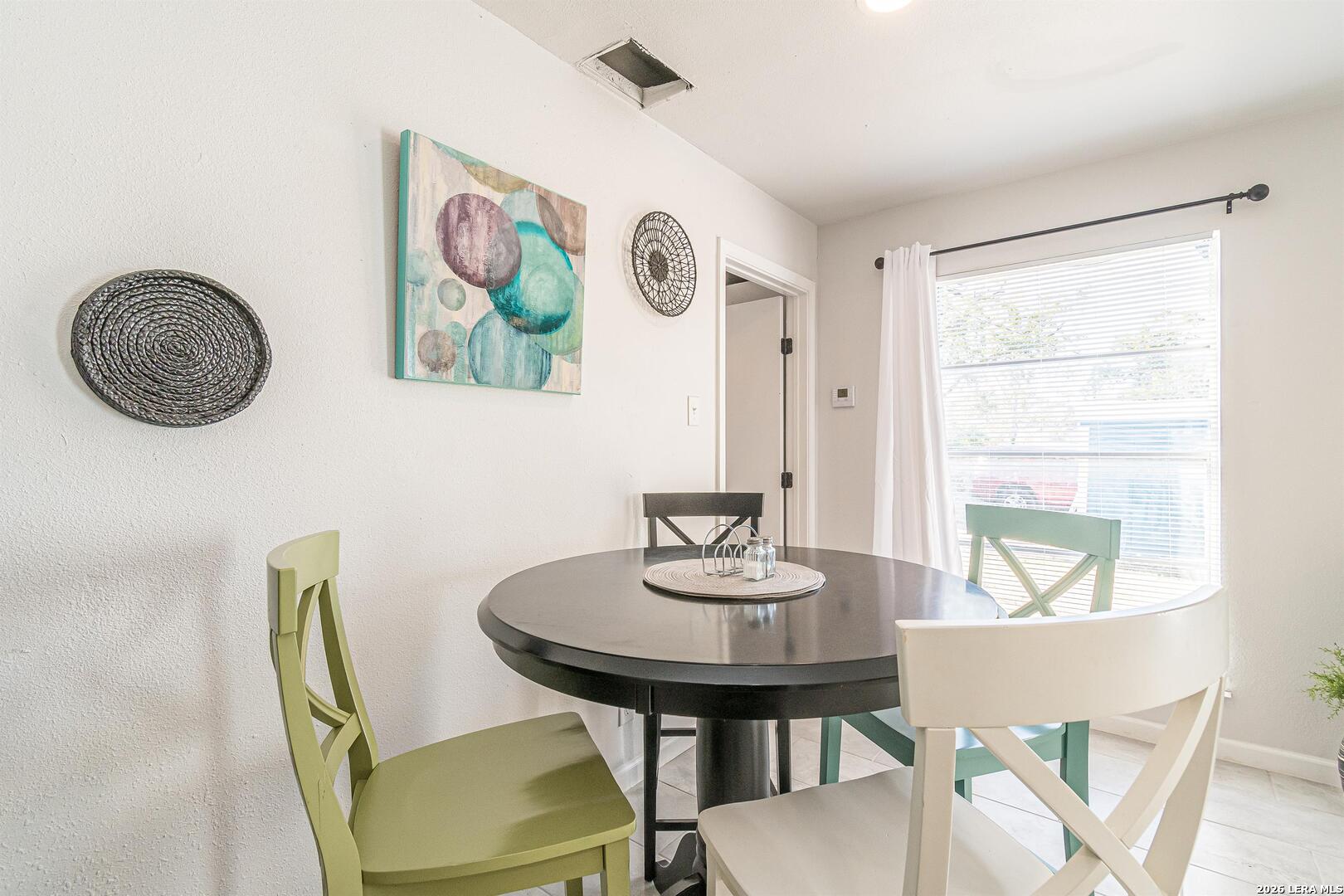 Dining area with a round table, colorful chairs, and a large window with blinds.