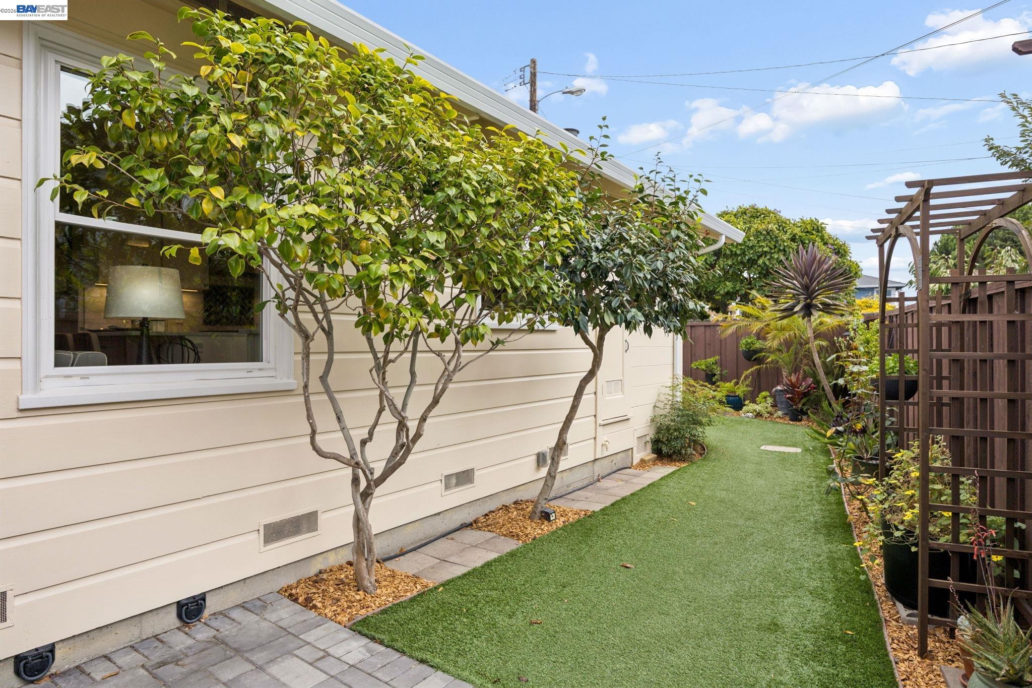 Landscaped side yard with a narrow lawn, mature trees, and a wooden pergola beside the house.