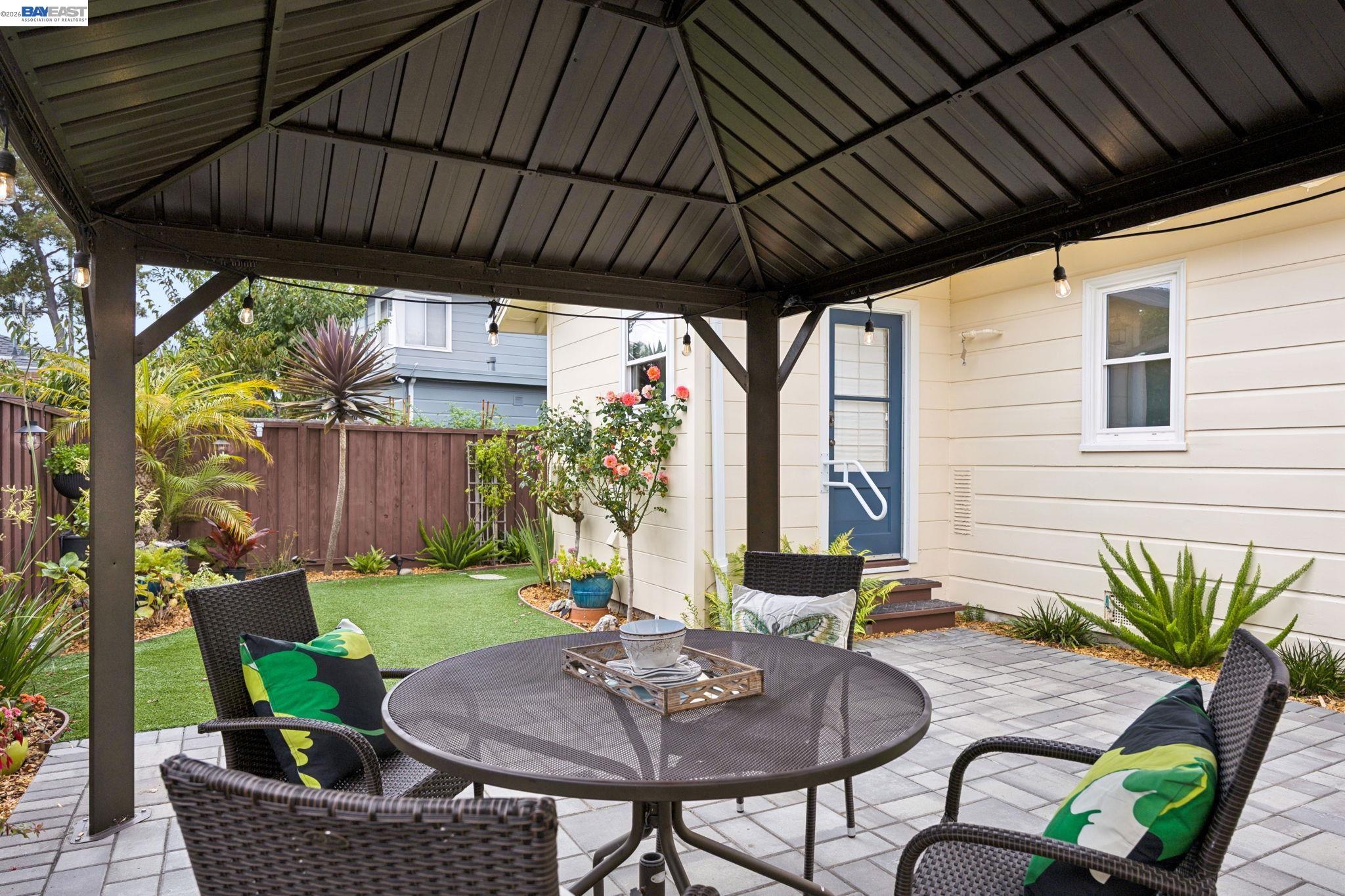 Covered backyard patio with wicker dining set, string lights, paved seating area, and landscaped garden.