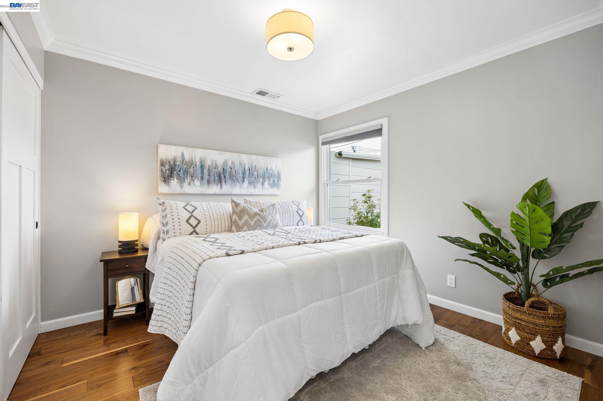 Staged bedroom with a white bed, hardwood floors, and a large potted plant beside the window.