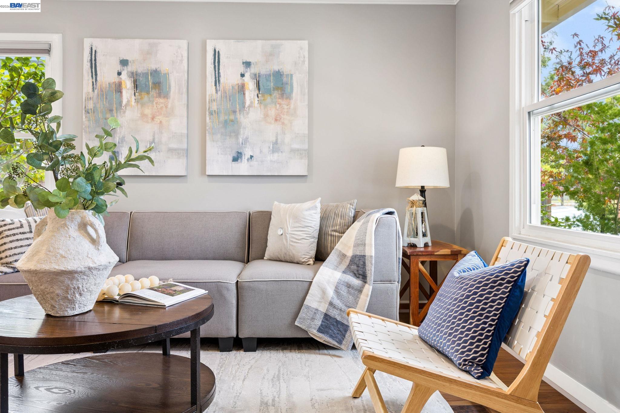 Living room with a gray sofa, abstract artwork, and a wooden lounge chair beside large windows.