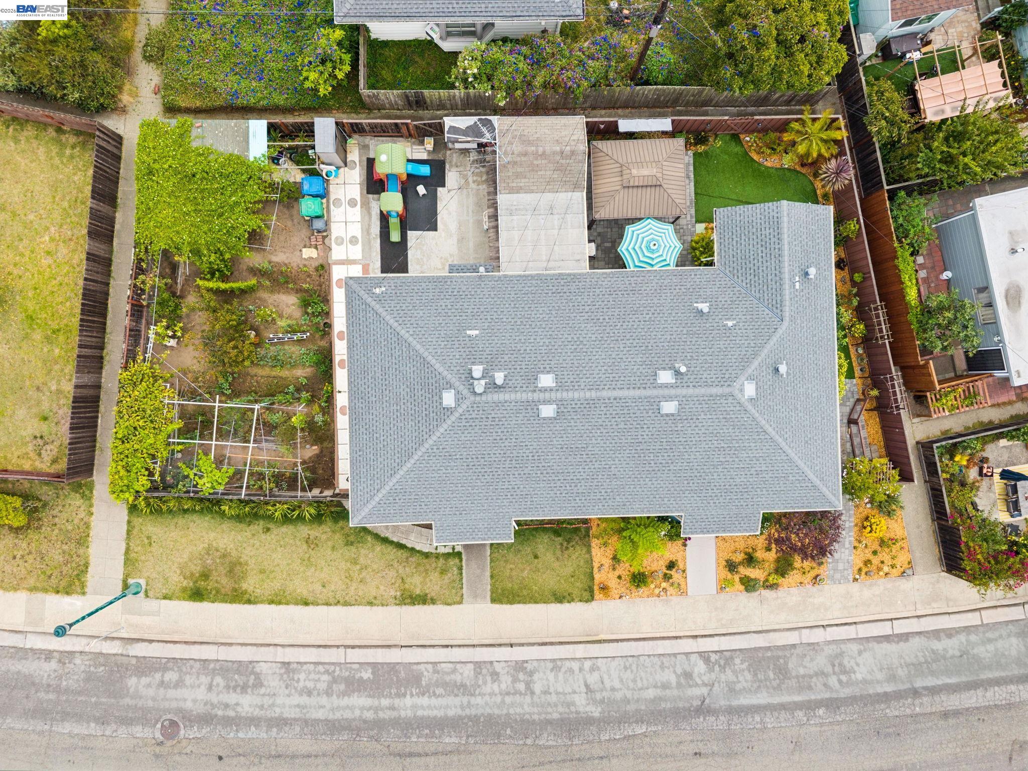 Aerial view of a home with a large gray roof, fenced garden, and backyard play area.