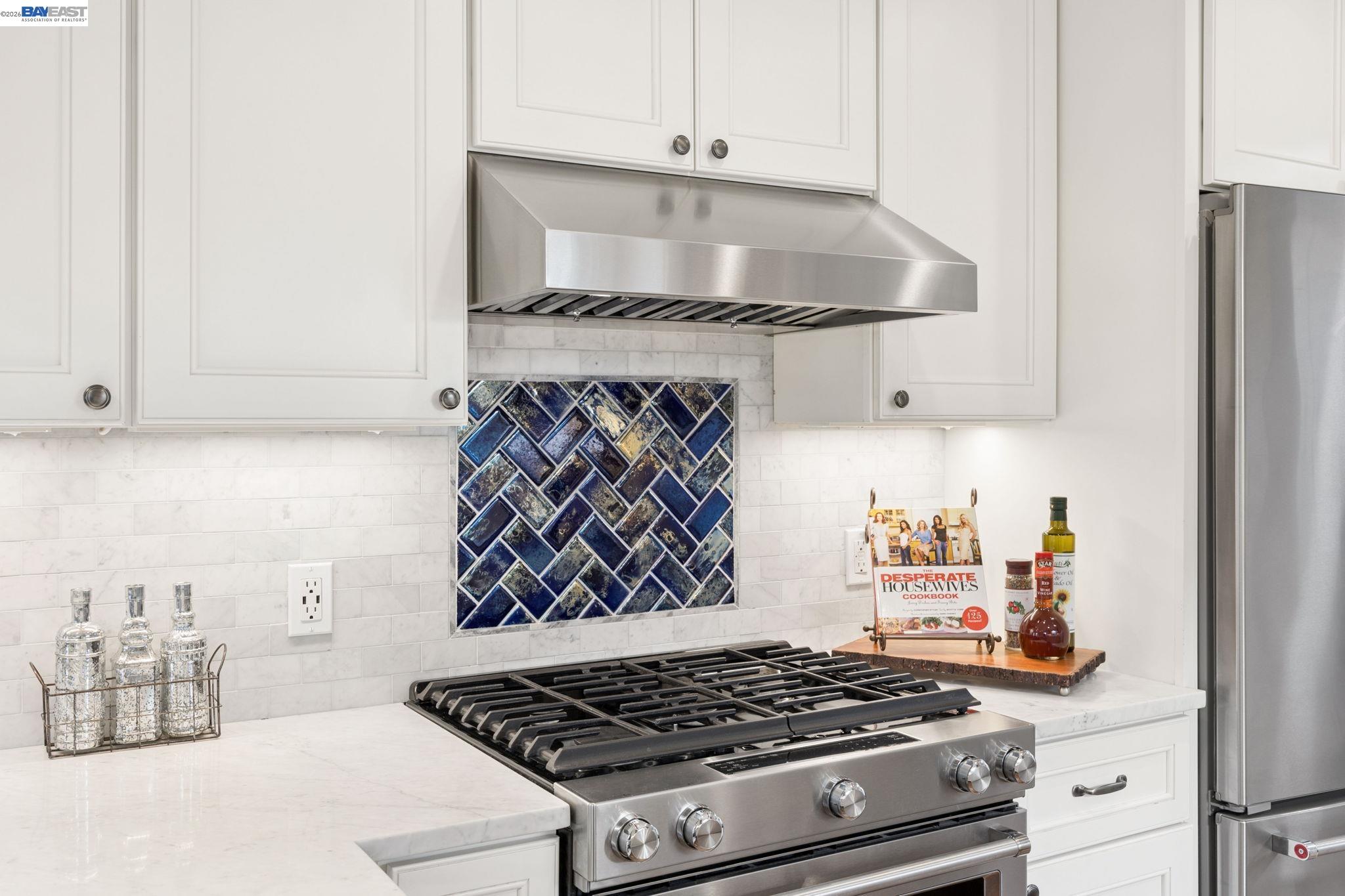 Kitchen range and stainless hood beneath blue patterned tile, surrounded by white cabinetry and marble counters.