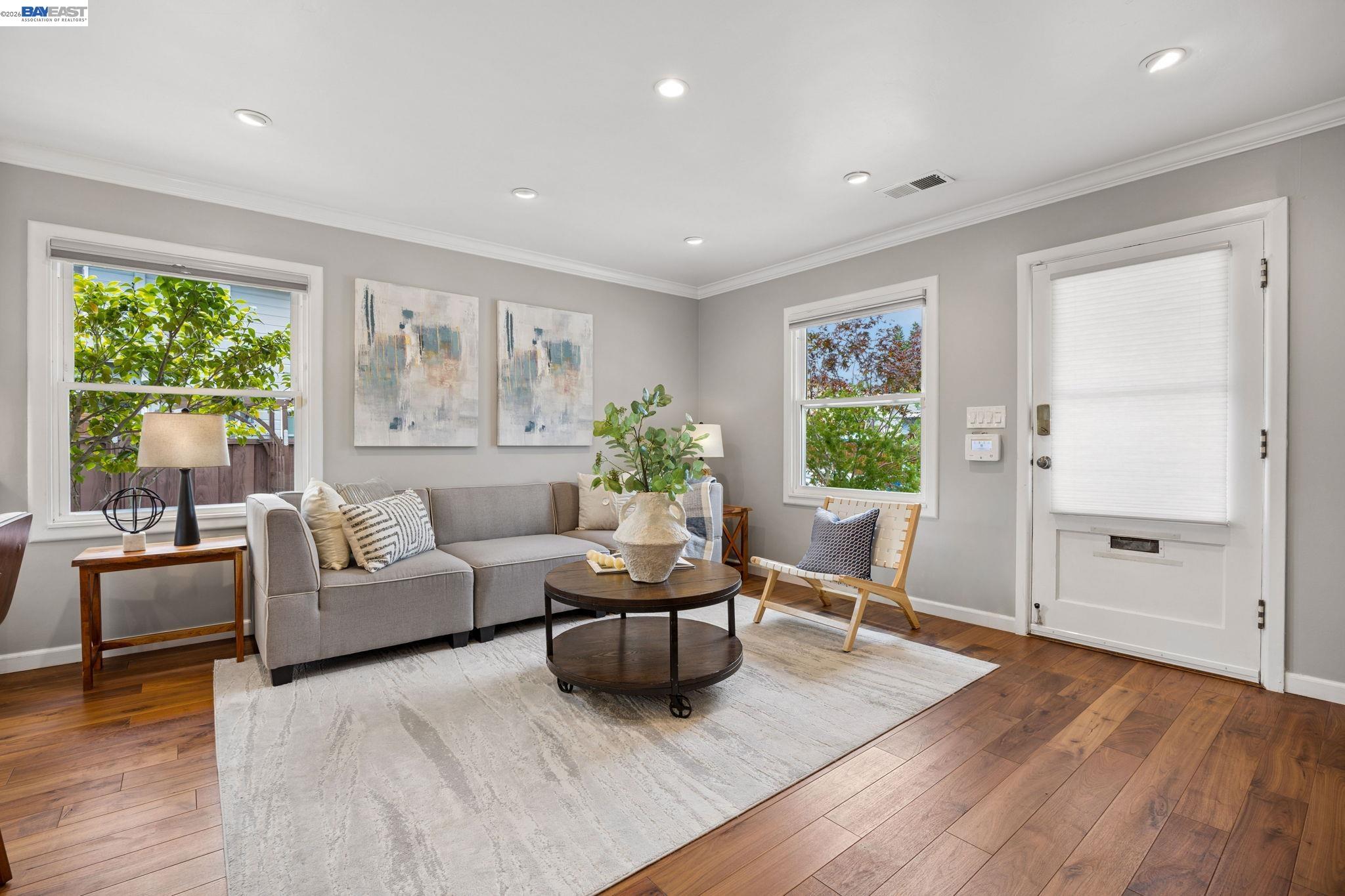 Staged living room with a gray sofa, round coffee table, and leafy views through two windows.