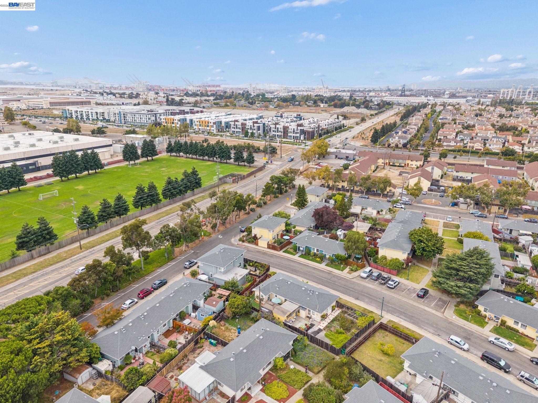 Aerial neighborhood view with homes, a soccer field, newer apartments, and port cranes on the horizon.